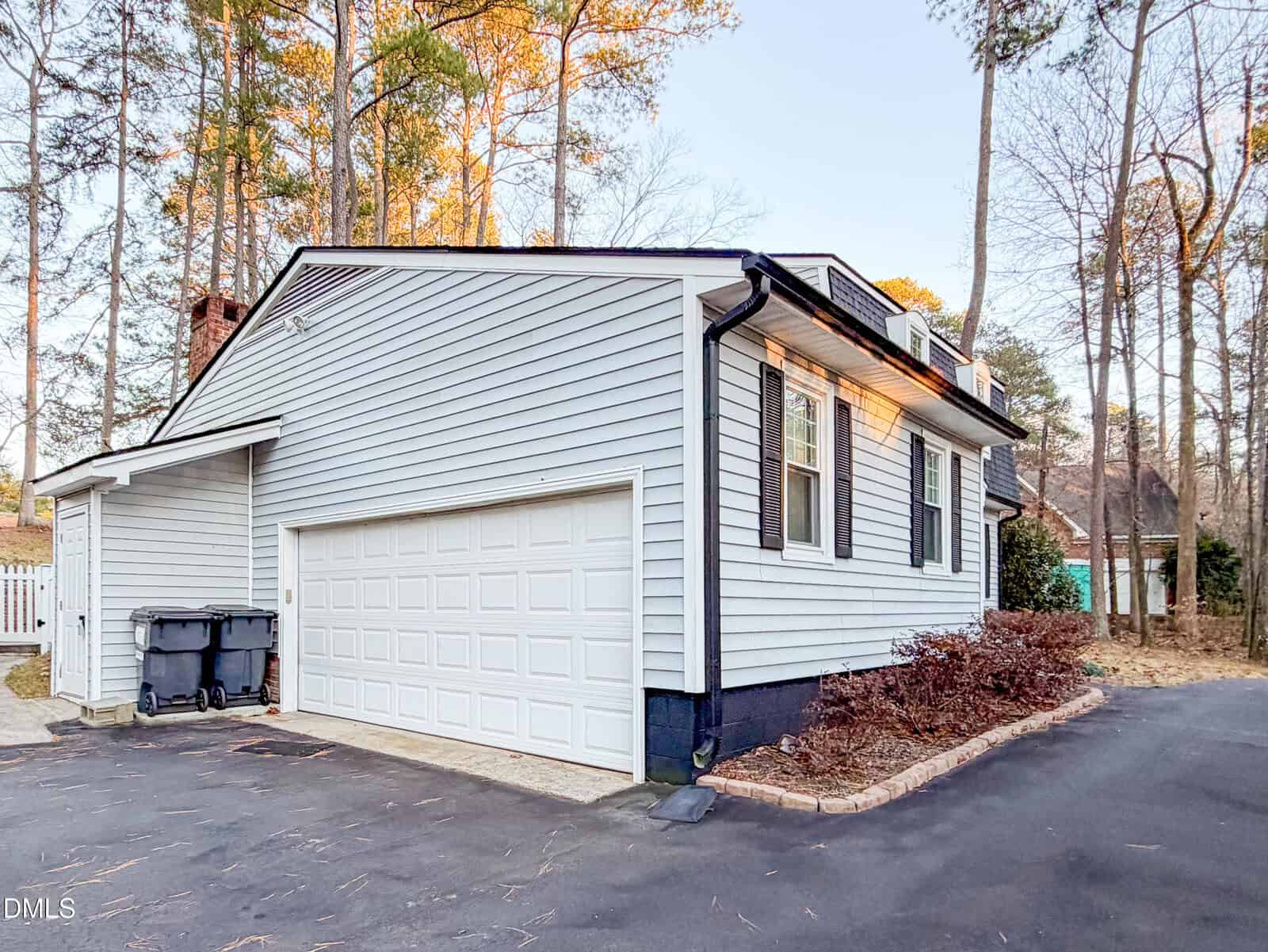 A light gray house with black shutters and a white double garage door sits next to a paved driveway. Trash bins are by the garage, trees surround the house, and landscaping lines the foundation.