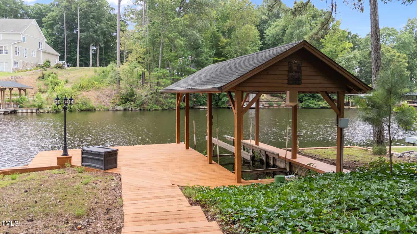 Wooden dock with a covered boat slip extends over a calm lake, surrounded by trees. A house is visible on a hill in the background. The dock has stairs, a lamp post, and a storage box. Sky is partly cloudy.