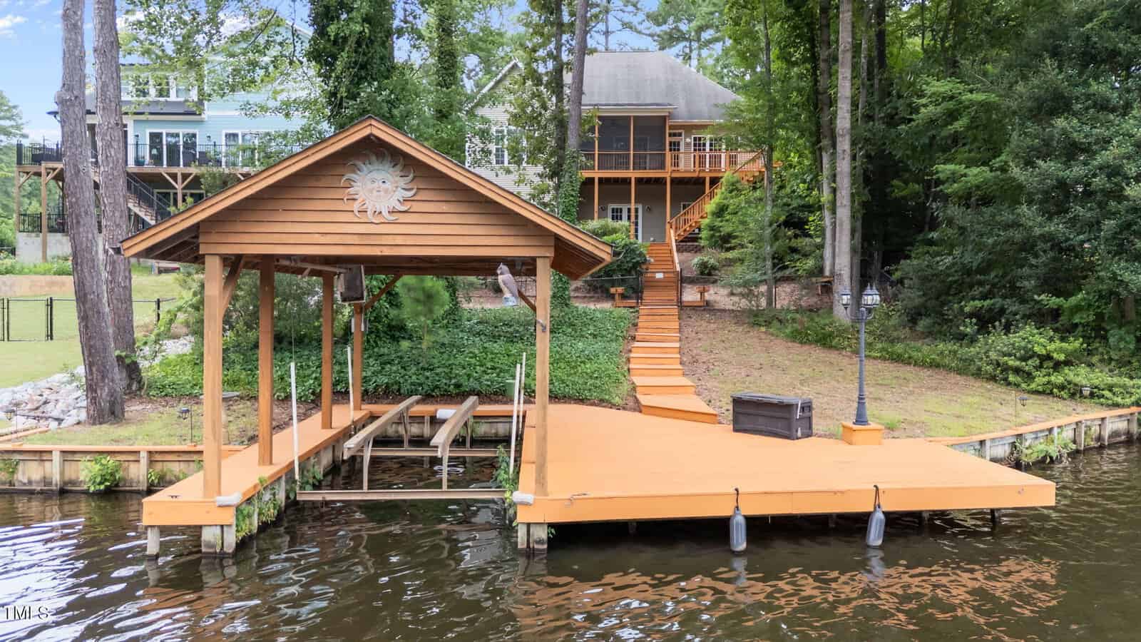 Wooden dock with a covered boat slip and stairs leading up to a two-story house surrounded by trees. The dock has a decorative sun emblem, bench, and lamp post, and the house features a large porch and upper balcony.
