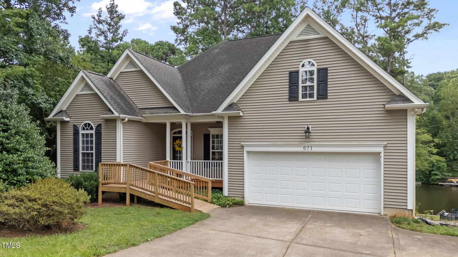 A single-story beige house with white trim, black shutters, a two-car garage, a wooden accessibility ramp, and a covered front porch awaits you at 671 Sagamore Drive. Nestled in Louisburg and surrounded by trees and a grassy lawn, this charming home offers both comfort and accessibility in a tranquil setting.