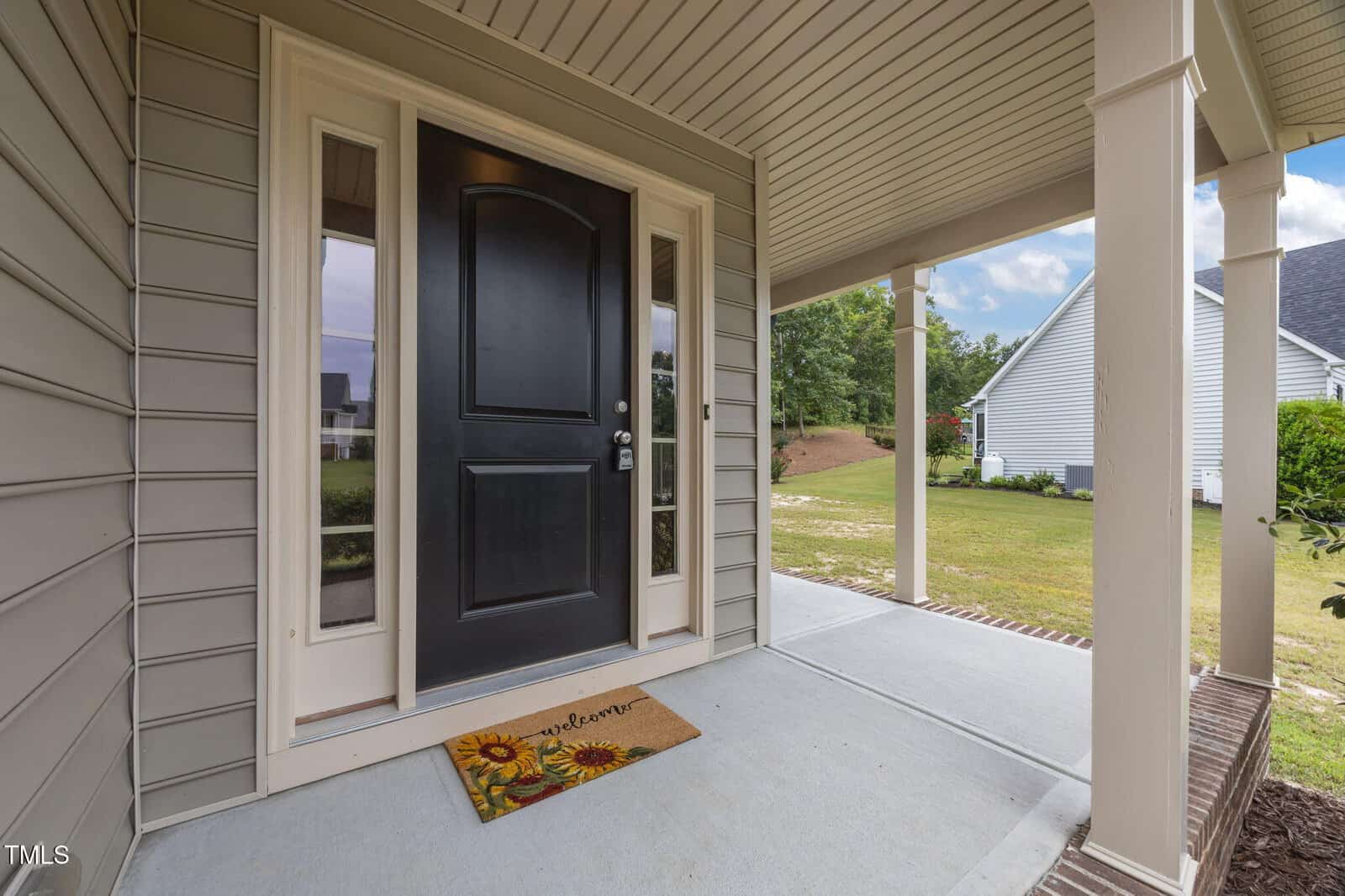 A covered front porch with beige siding and a black door, flanked by windows. A doormat with sunflowers and "welcome" written on it lies in front of the door. The porch overlooks a grassy yard and neighboring homes.