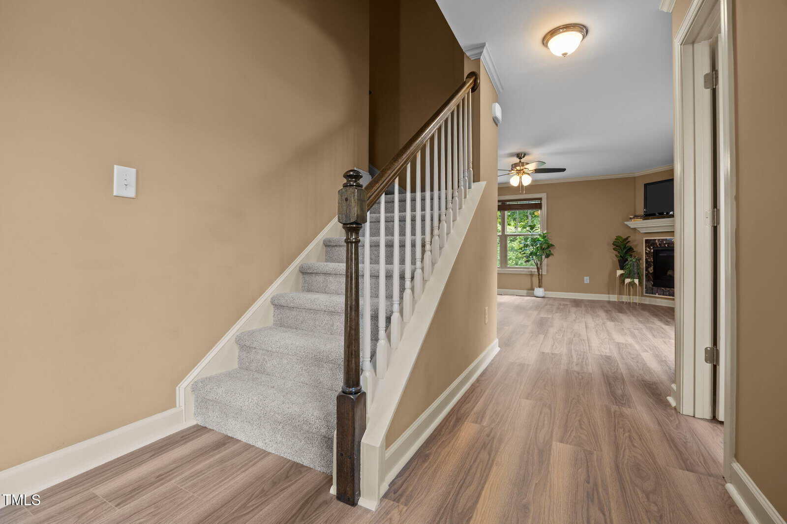 A staircase with carpeted steps and a wooden handrail beside a hallway with light wood flooring, leading to a living room with a fireplace, ceiling fan, and large window.