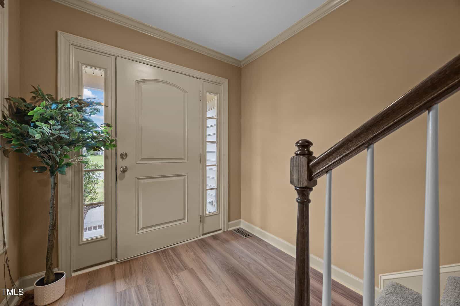 A beige entryway features a front door with two narrow windows on either side, a potted artificial plant on the left, and a staircase with a dark wood handrail and white spindles on the right.