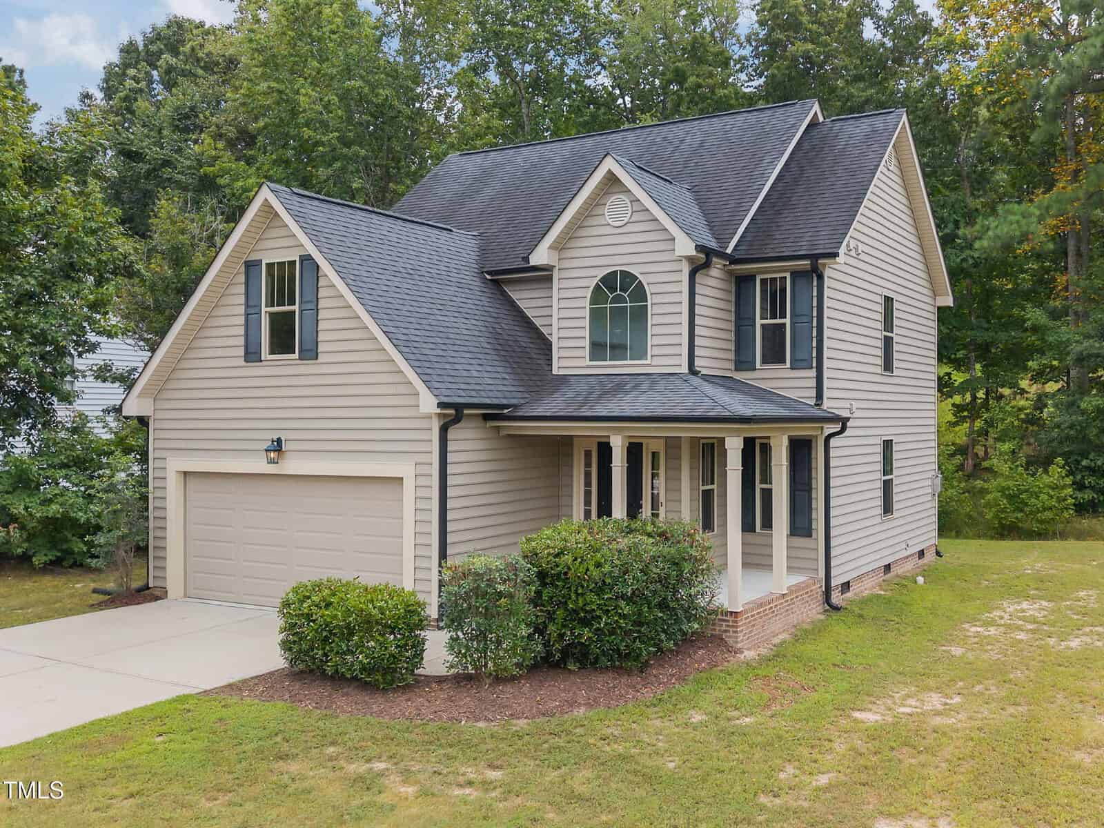 Two-story beige house with black roof, attached two-car garage, covered front porch, and neatly trimmed bushes in front. Surrounded by trees and greenery with a concrete driveway leading to the garage.