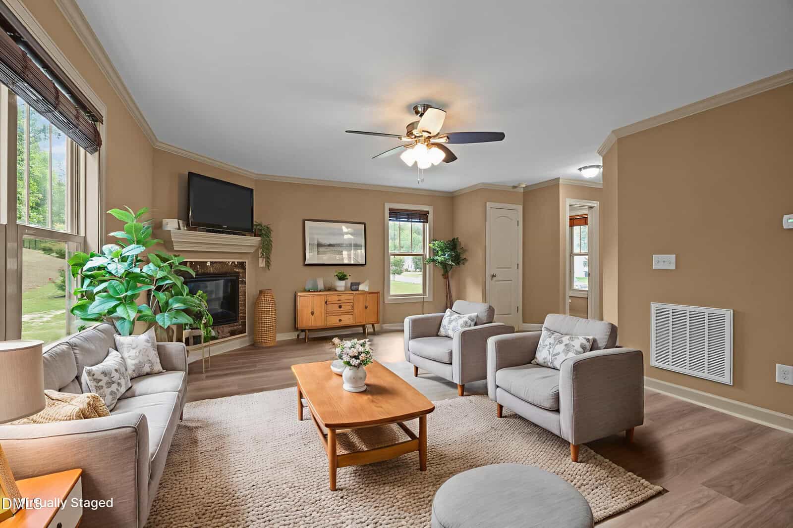 A living room with beige walls, light wood flooring, and a ceiling fan. The furniture includes a gray sofa, two gray armchairs, a wooden coffee table, and a sideboard, with plants and framed art for decor.