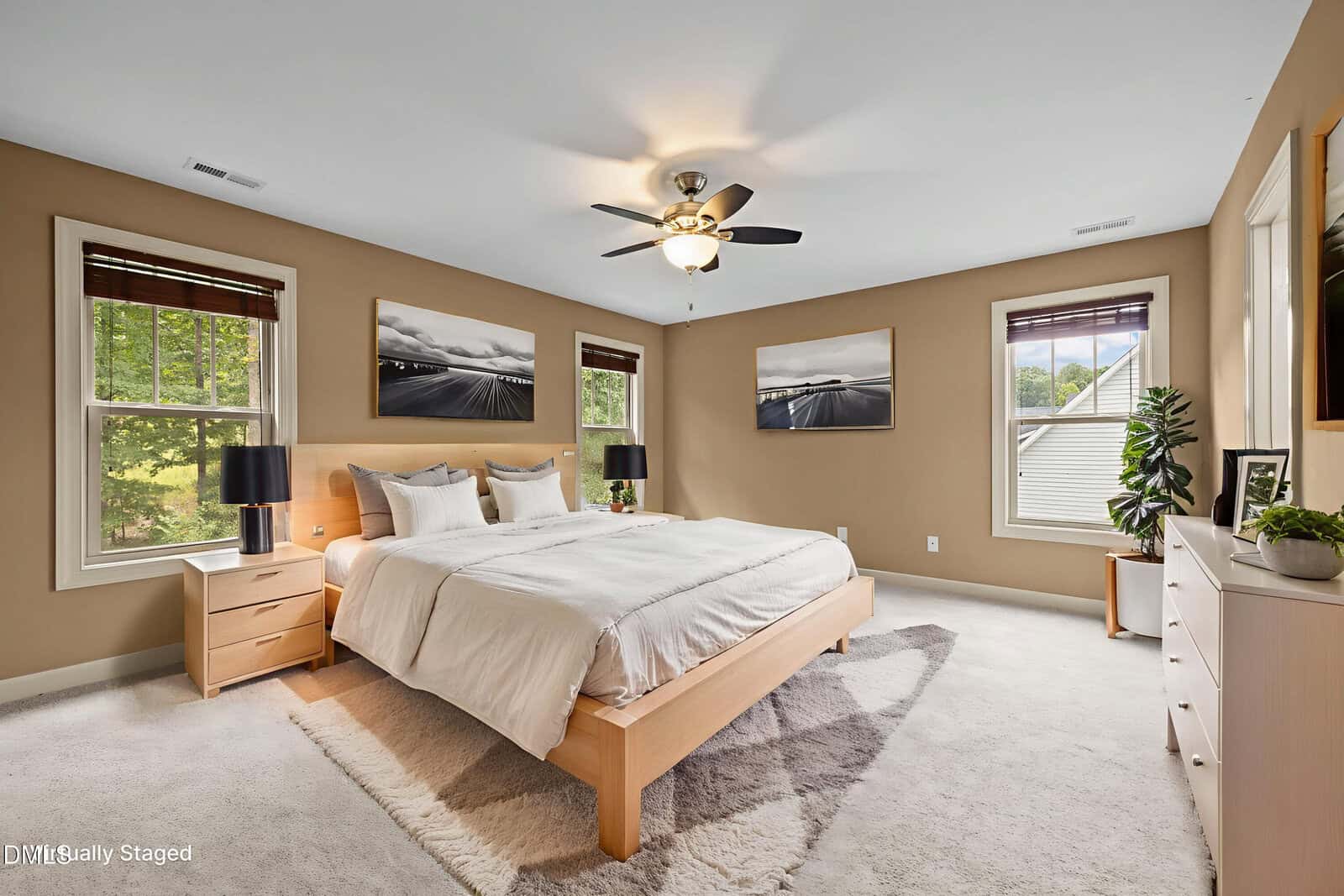 A spacious bedroom with beige walls, a large bed with white bedding, two nightstands with lamps, three landscape photos on the wall, carpeted floor, ceiling fan, and multiple windows letting in natural light.