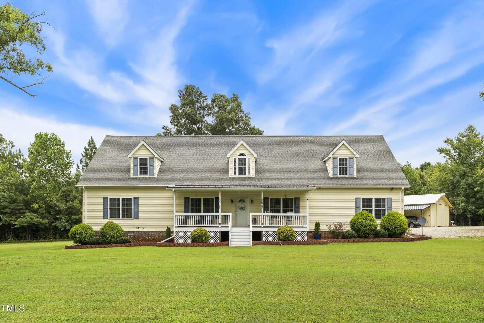 A yellow two-story house with dormer windows, a covered front porch, and a neatly trimmed lawn, surrounded by trees under a partly cloudy blue sky.