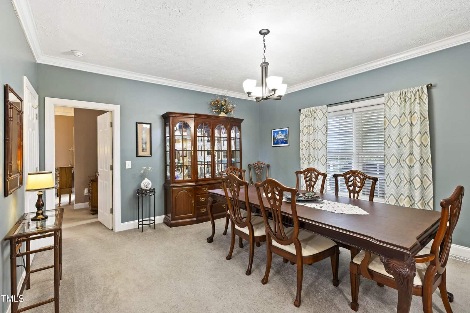 Formal dining room with a long wooden table, eight chairs, a china cabinet, chandelier, and decorative items. The walls are painted blue-gray, and there are two windows with patterned curtains. Carpeted floor.