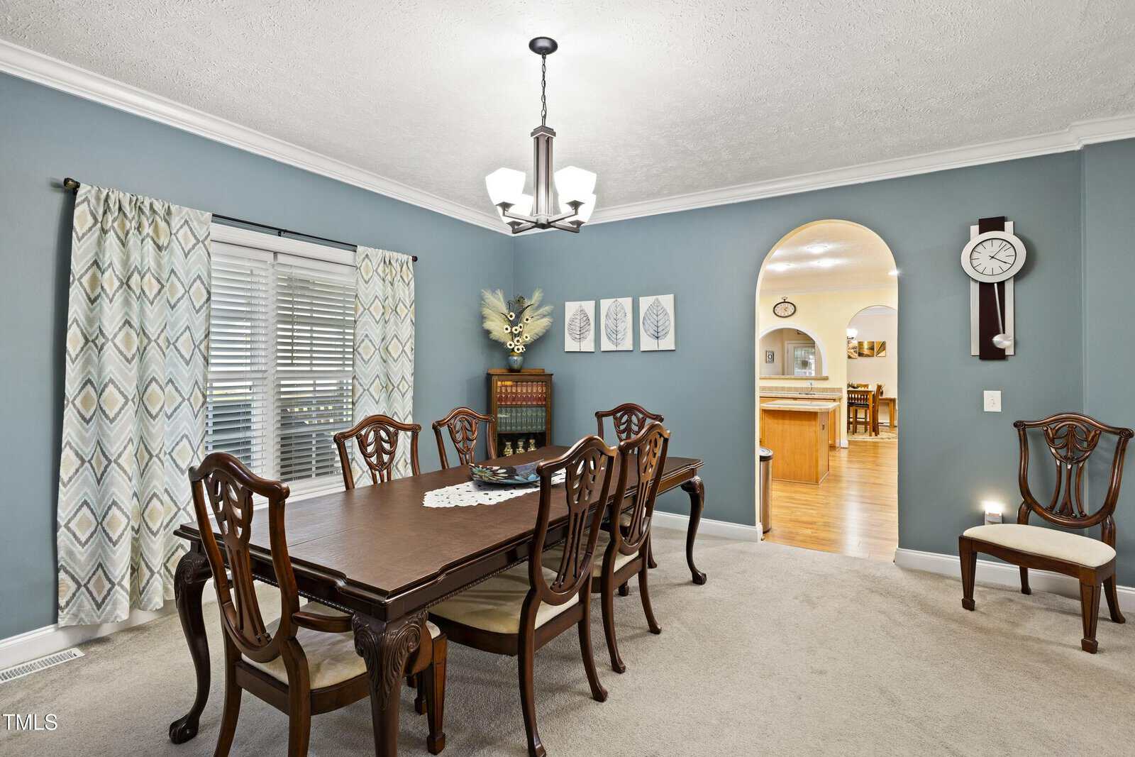 A dining room with a dark wooden table and eight chairs, light blue walls, patterned curtains on a window, artwork and a clock on the wall, and an archway leading to a kitchen area with wood flooring.