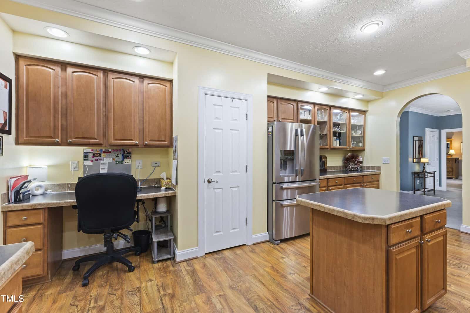 A kitchen with light yellow walls, brown cabinets, stainless steel appliances, a center island, a built-in desk area with a black chair, and wood flooring. A door is centered on the back wall.