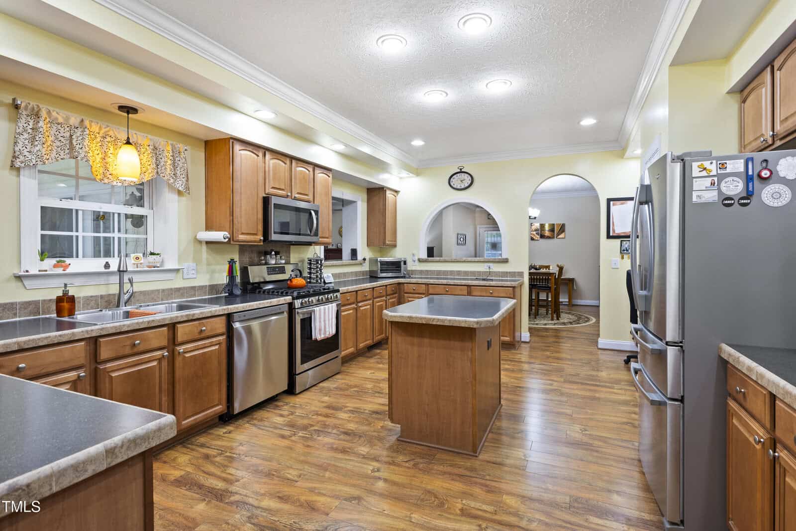 Spacious kitchen with wood cabinets, stainless steel appliances, a central island, dark countertops, and wood flooring. A window above the sink and bright ceiling lights provide ample illumination. Dining area visible through an archway.