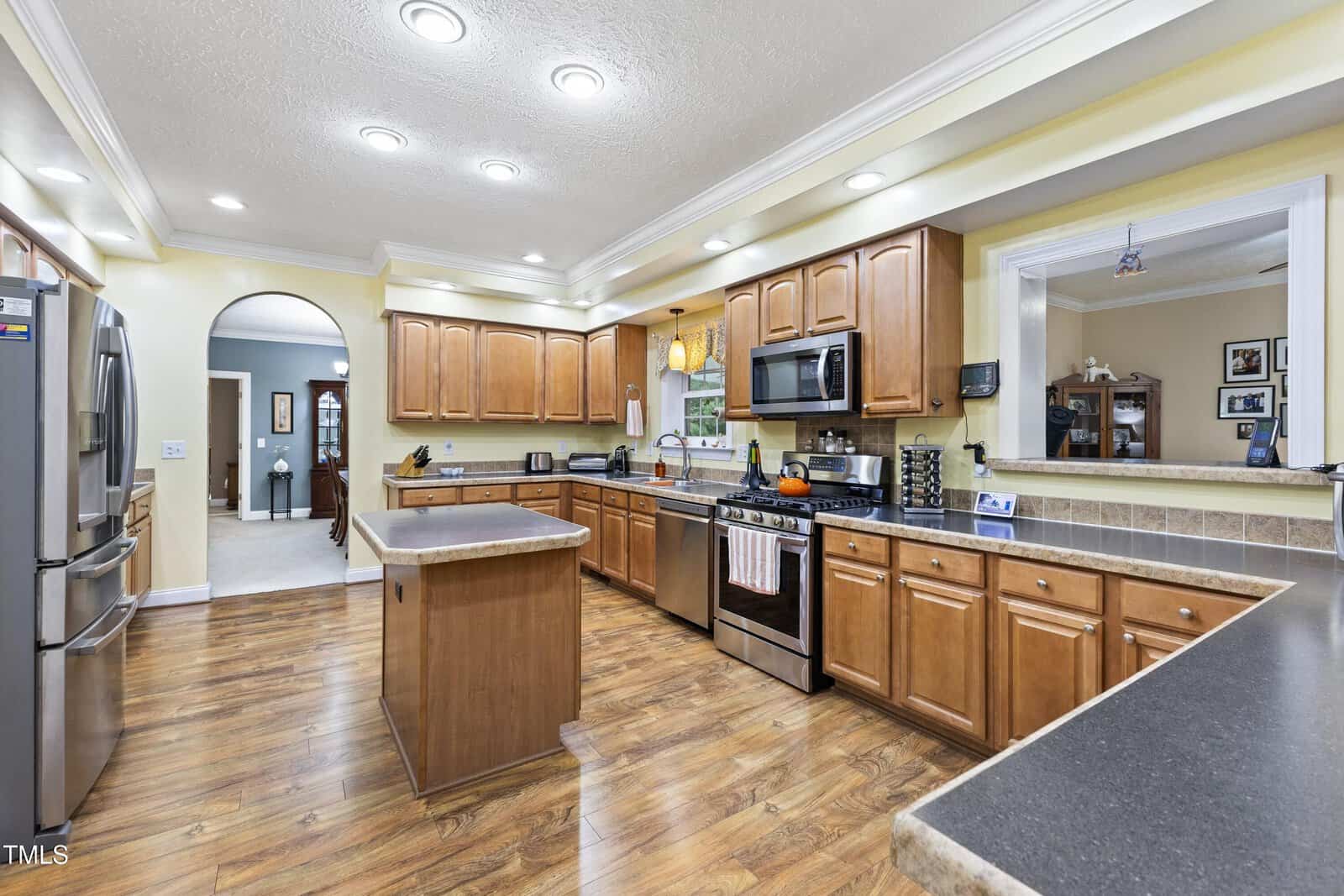 Spacious kitchen with wood cabinets, stainless steel appliances, an island in the center, hardwood floors, and recessed lighting. A window is above the sink, and an archway leads to another room.