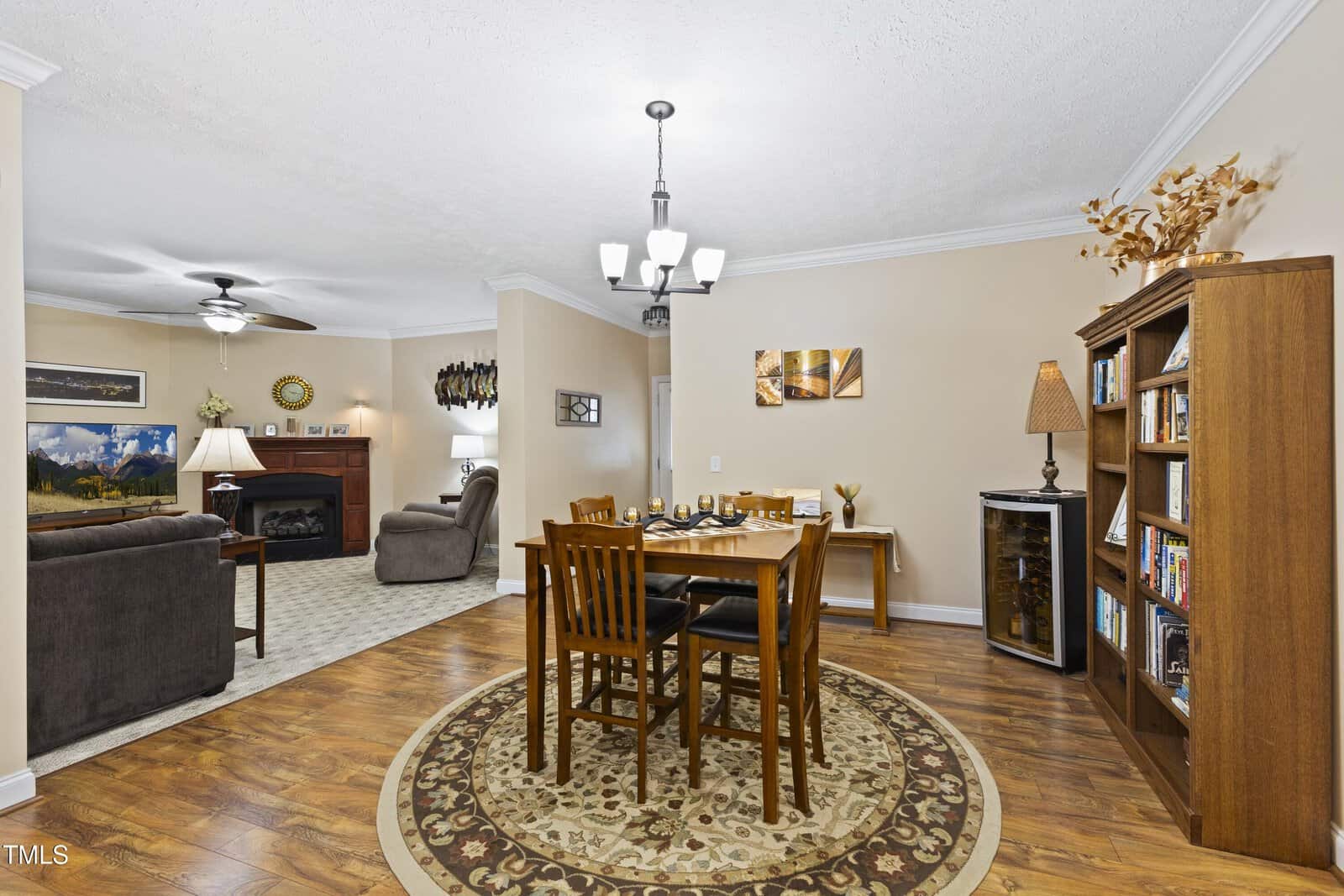 A cozy living and dining area with a round rug under a wooden table, bookshelves, a lamp, sofas, a fireplace, wall art, and a ceiling fan. Hardwood floors and neutral walls create a warm, inviting atmosphere.