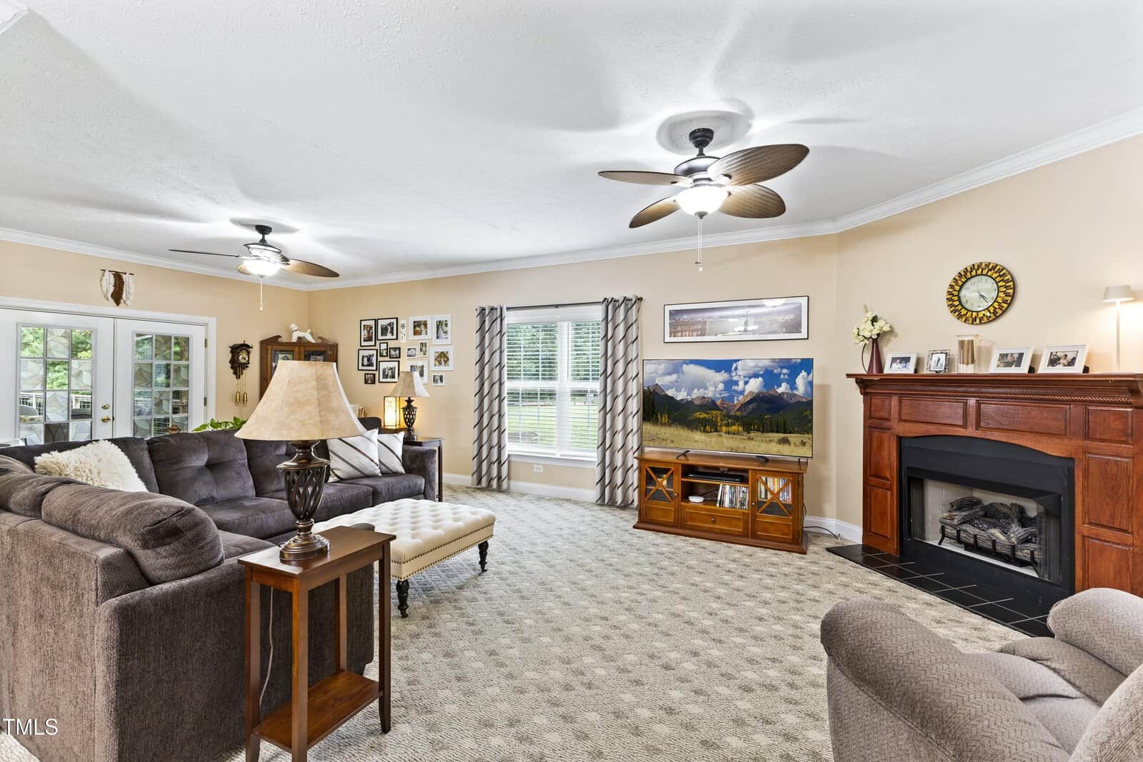 Spacious living room with beige walls, two ceiling fans, brown sofas, fireplace, TV, and large windows with patterned curtains. Family photos decorate the walls.