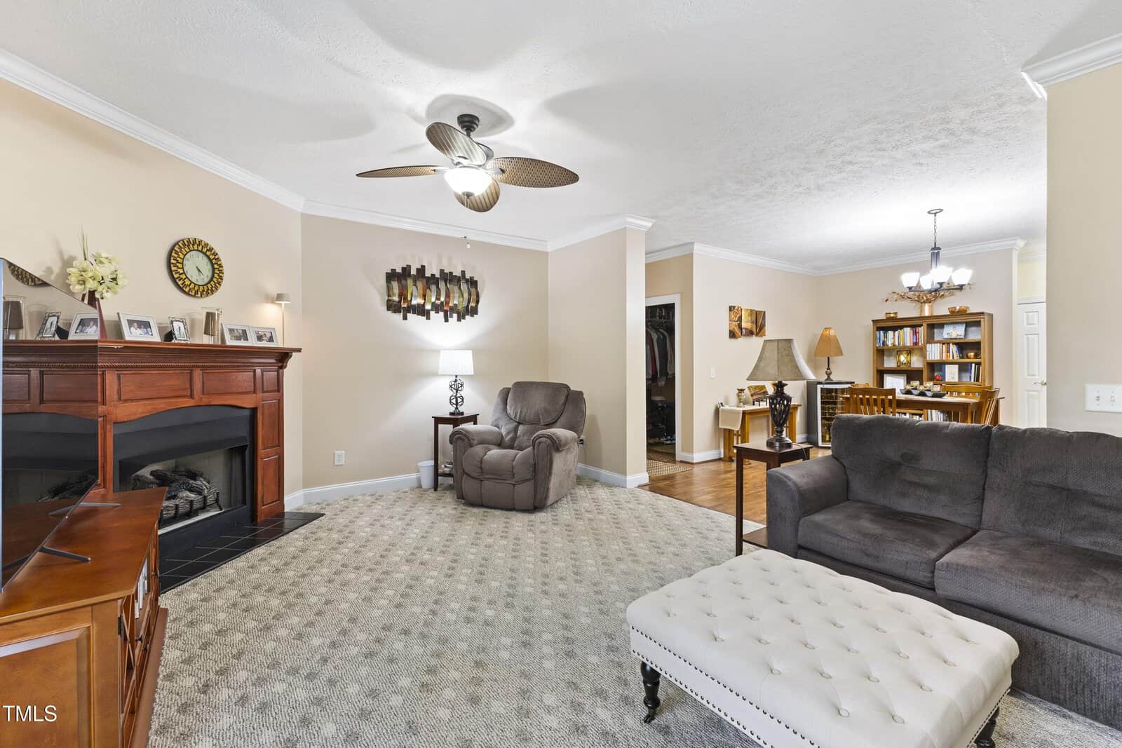 A cozy living room with a fireplace, armchair, sofa, ottoman, ceiling fan, and carpet. Photos and decor adorn the mantel; a dining area and bookshelf are visible in the background.
