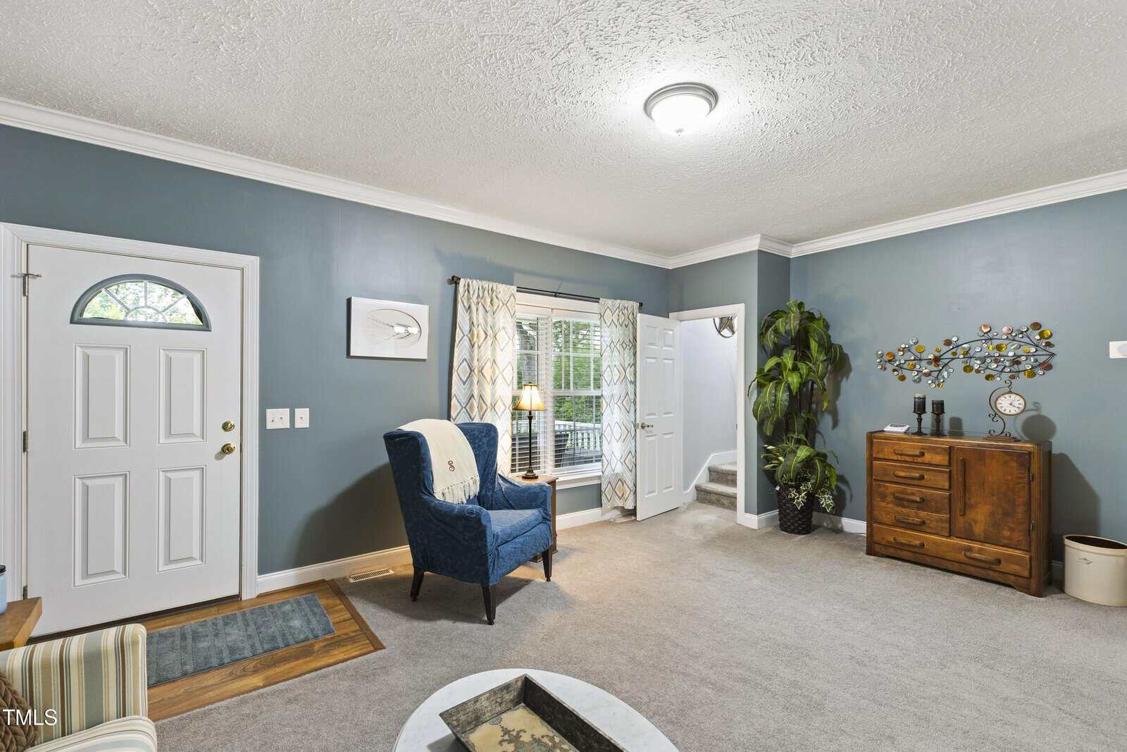A living room with blue-gray walls, white trim, and a textured ceiling. It features a blue armchair, a wooden dresser, a potted plant, wall art, and a window with patterned curtains next to a white door.