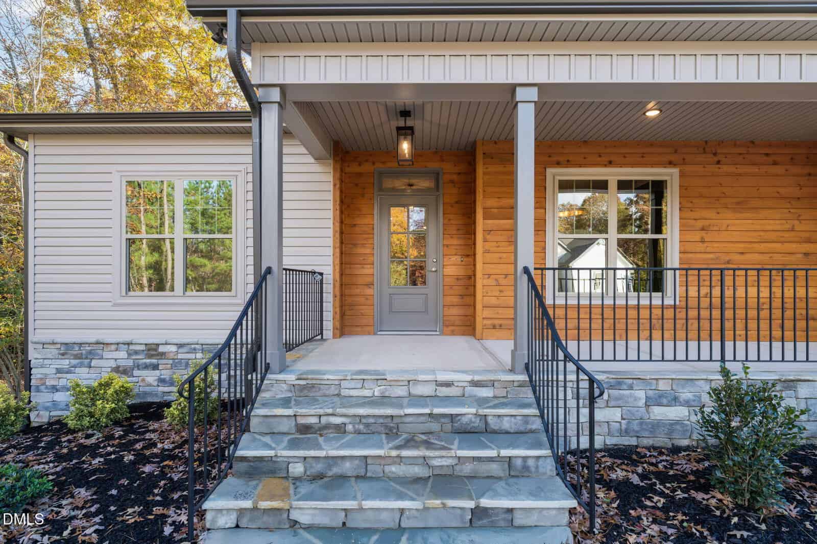 Front porch of a modern house with stone steps, metal railings, a gray door, large windows, wood and light-colored siding, and landscaping with bushes and mulch.