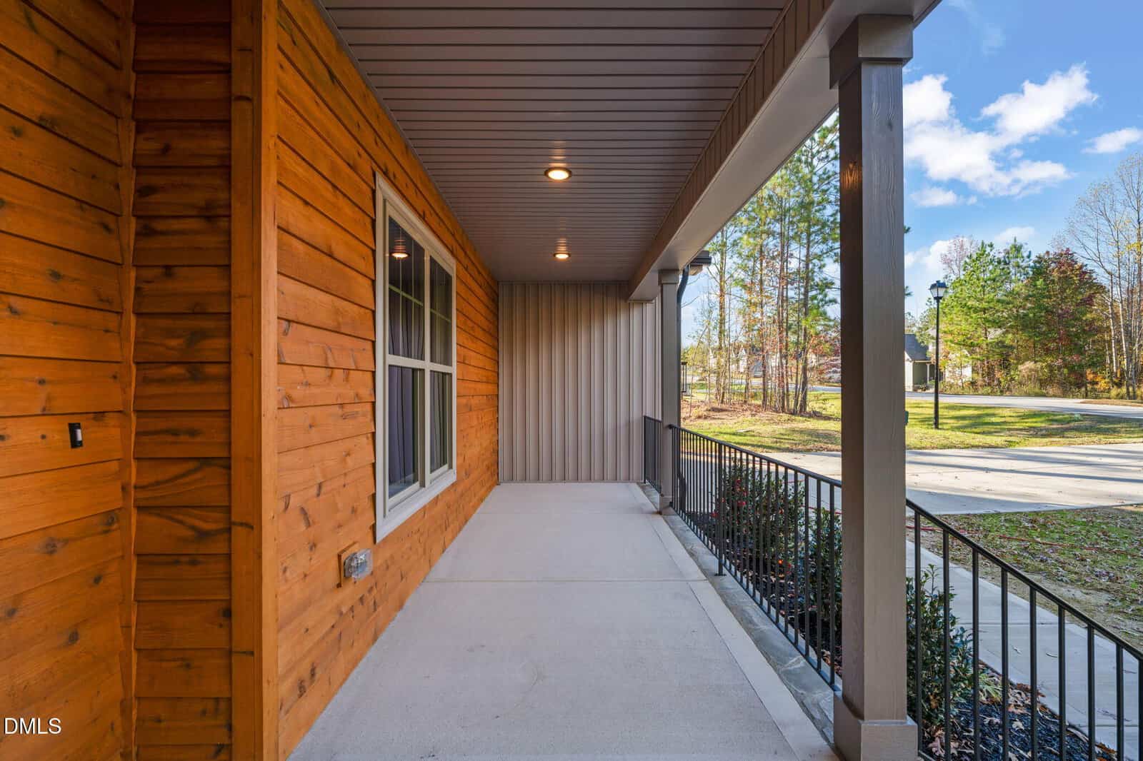 A covered front porch with wood siding, a concrete floor, metal railing, and a view of trees and a street in a suburban neighborhood.