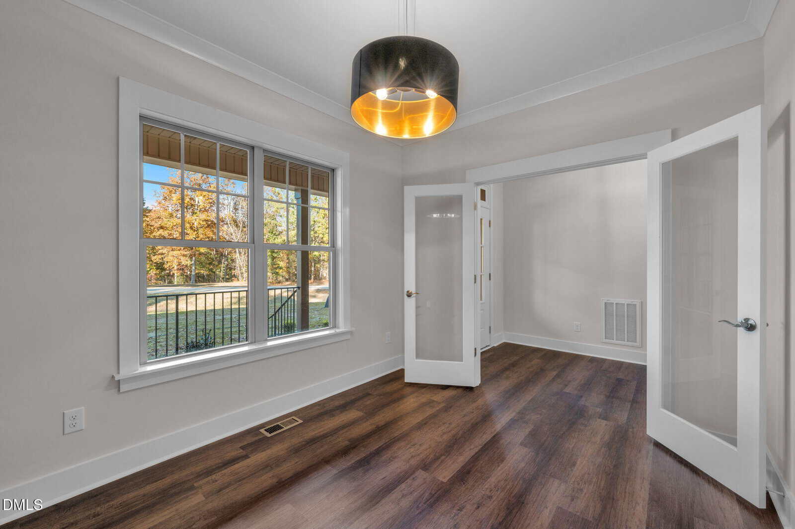 A bright room with wood floors, cream walls, a large window overlooking greenery, and double glass doors. A modern black and gold pendant light hangs from the ceiling.