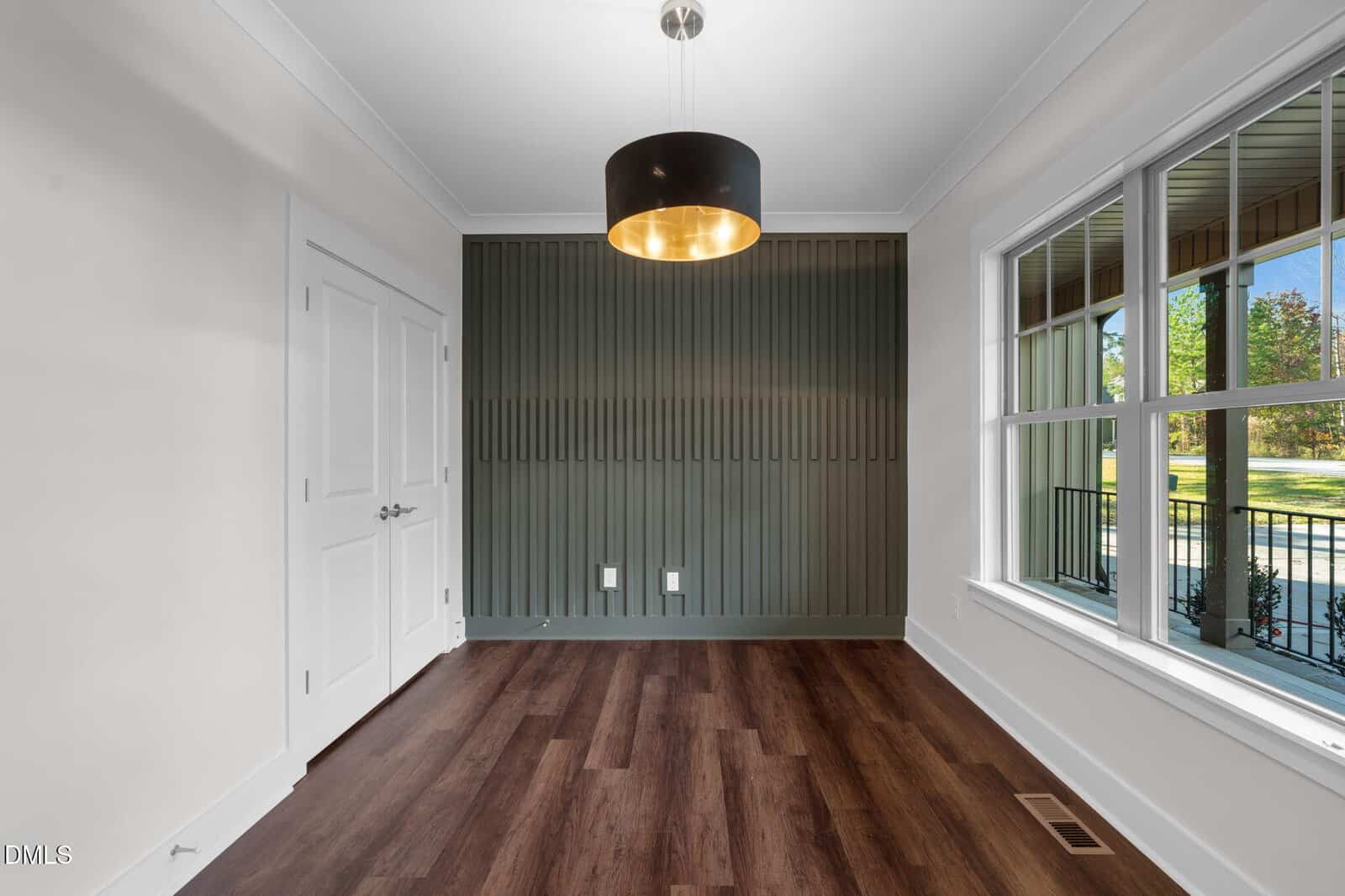 A modern empty room with dark wood flooring, white walls, a green accent wall with vertical grooves, a large window, double doors, and a black and gold ceiling light fixture.