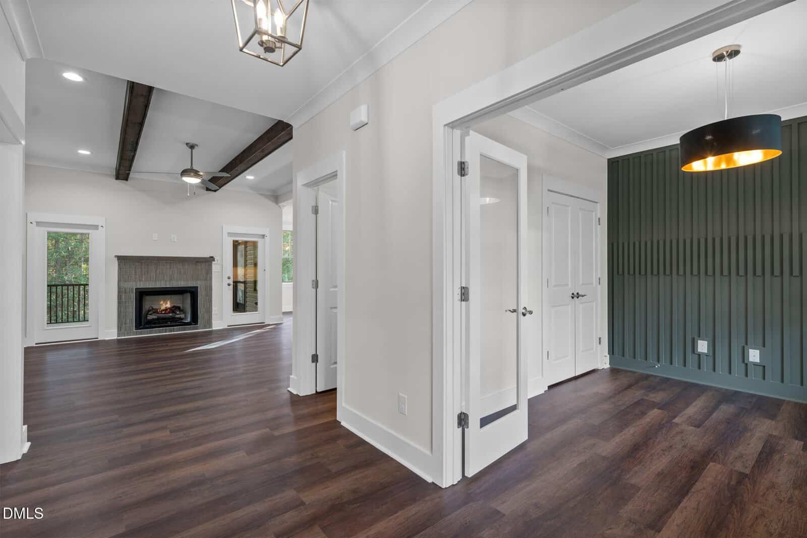Open-concept living area with dark wood floors, white walls, exposed ceiling beams, a fireplace, and an adjacent room with green accent wall and glass French doors. Large windows provide natural light.