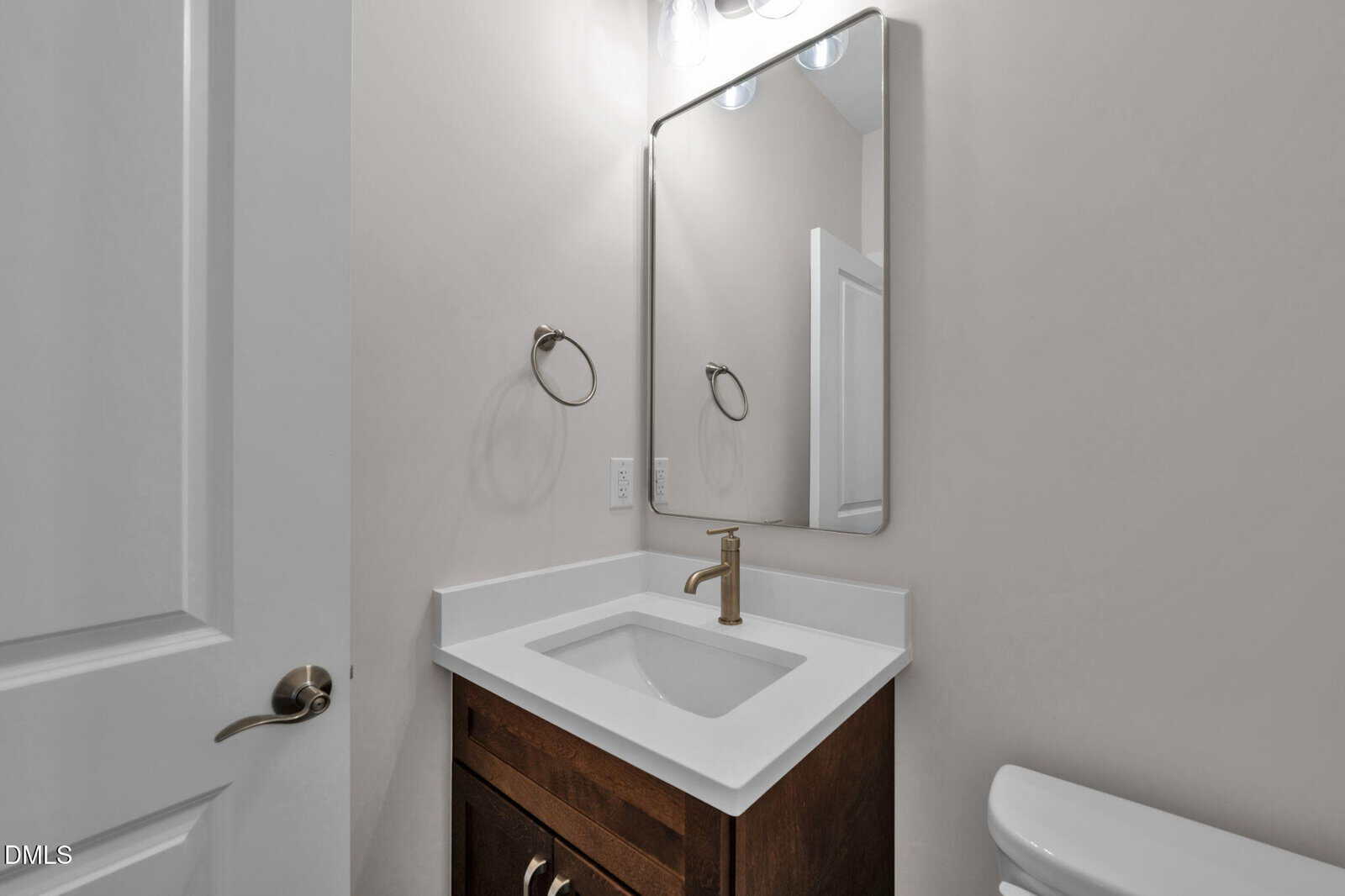 A small bathroom features a white countertop with an under-mount sink, a bronze faucet, a dark wood vanity, a wall-mounted mirror, towel ring, and part of a white toilet against light-colored walls.