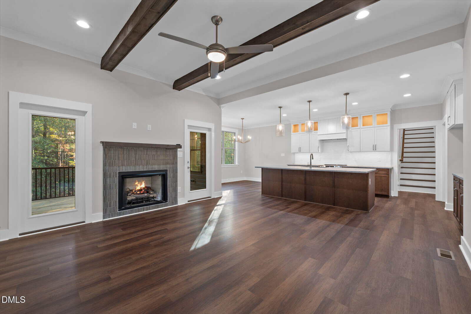 Open-concept living area with dark wood floors, a fireplace, ceiling fan, exposed beams, and large windows. The modern kitchen features an island, pendant lights, white cabinetry, and nearby stairs.