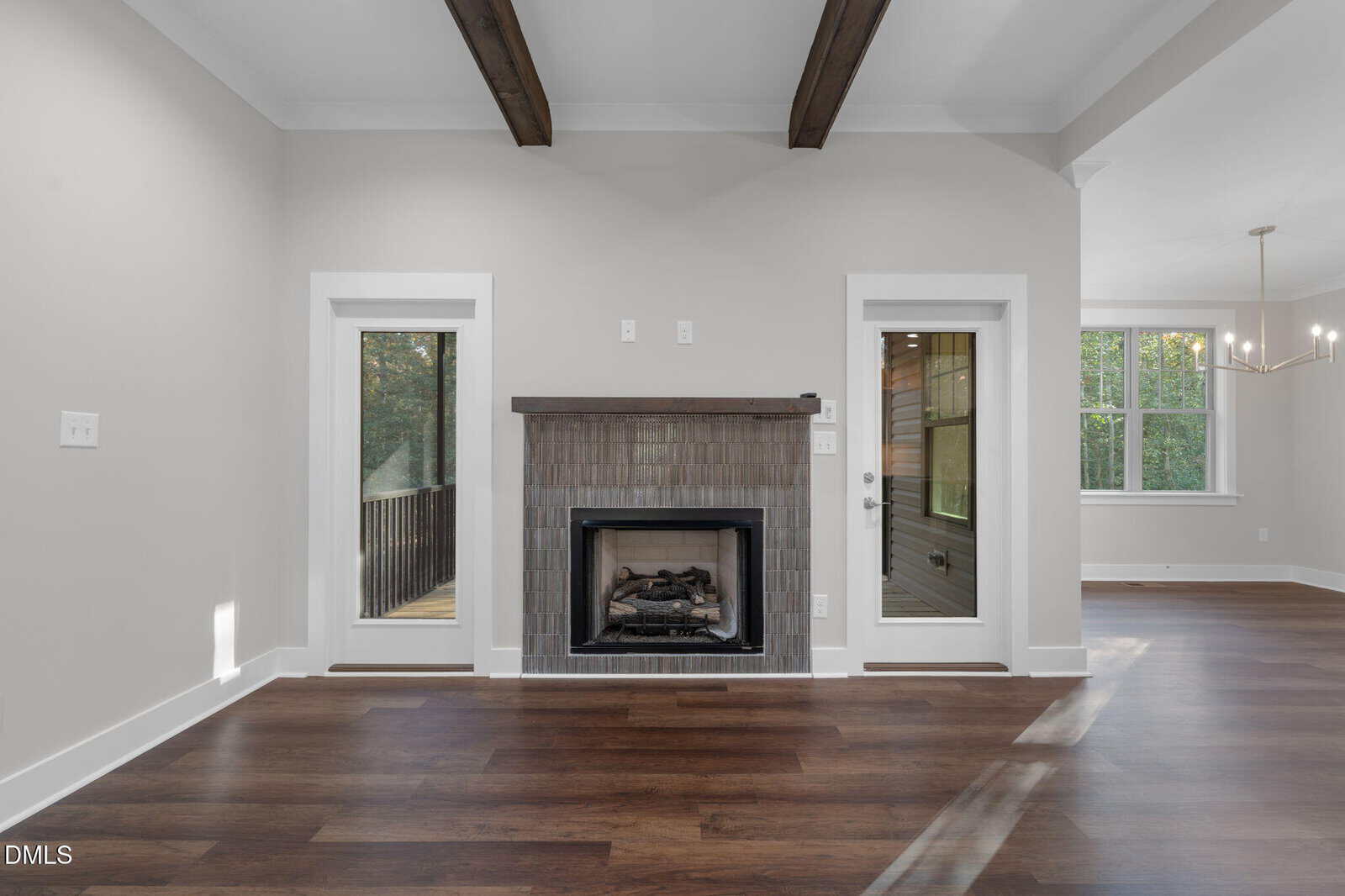 Living room with dark wood floors, ceiling beams, a gray tile fireplace, and two glass doors on either side. Sunlight streams in from an adjacent room with large windows and a modern chandelier.