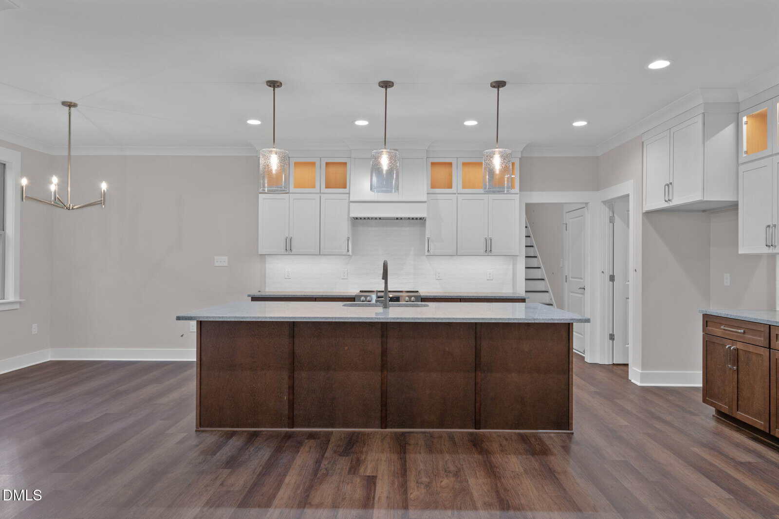 Modern kitchen with white cabinets, dark wood island, pendant lights, stainless steel appliances, and hardwood floors. Staircase and chandelier are visible in the background.