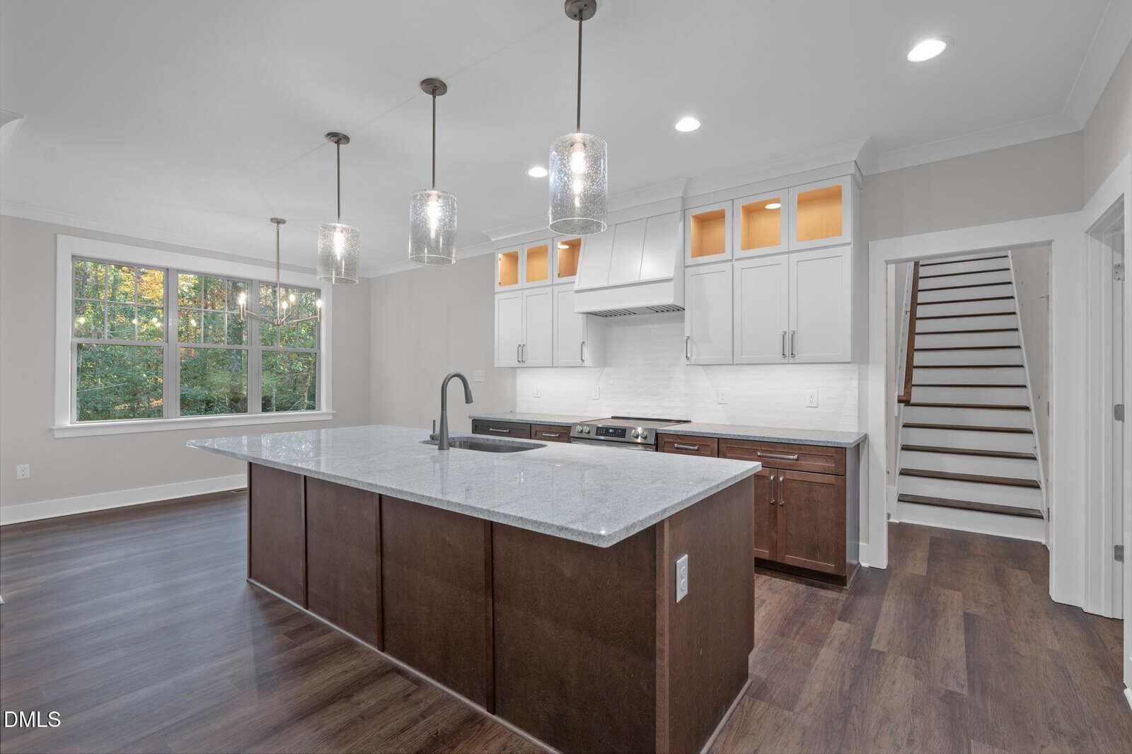 Modern kitchen with a large island, dark lower cabinets, white upper cabinets, stainless steel appliances, pendant lights, wood floors, and a staircase in the background. Large window lets in natural light.