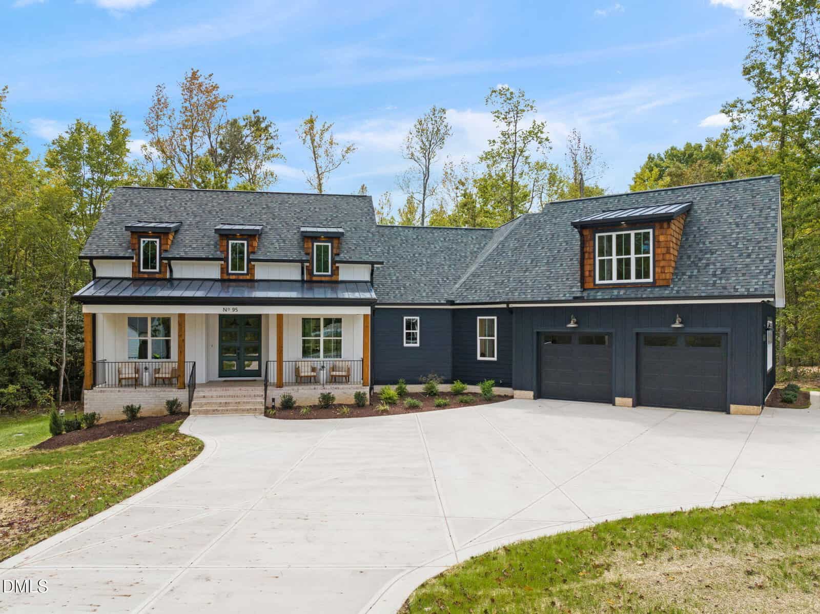 A modern blue house with white trim, a front porch, dormer windows, and two-car garage sits on a spacious concrete driveway, surrounded by trees and greenery under a clear blue sky.