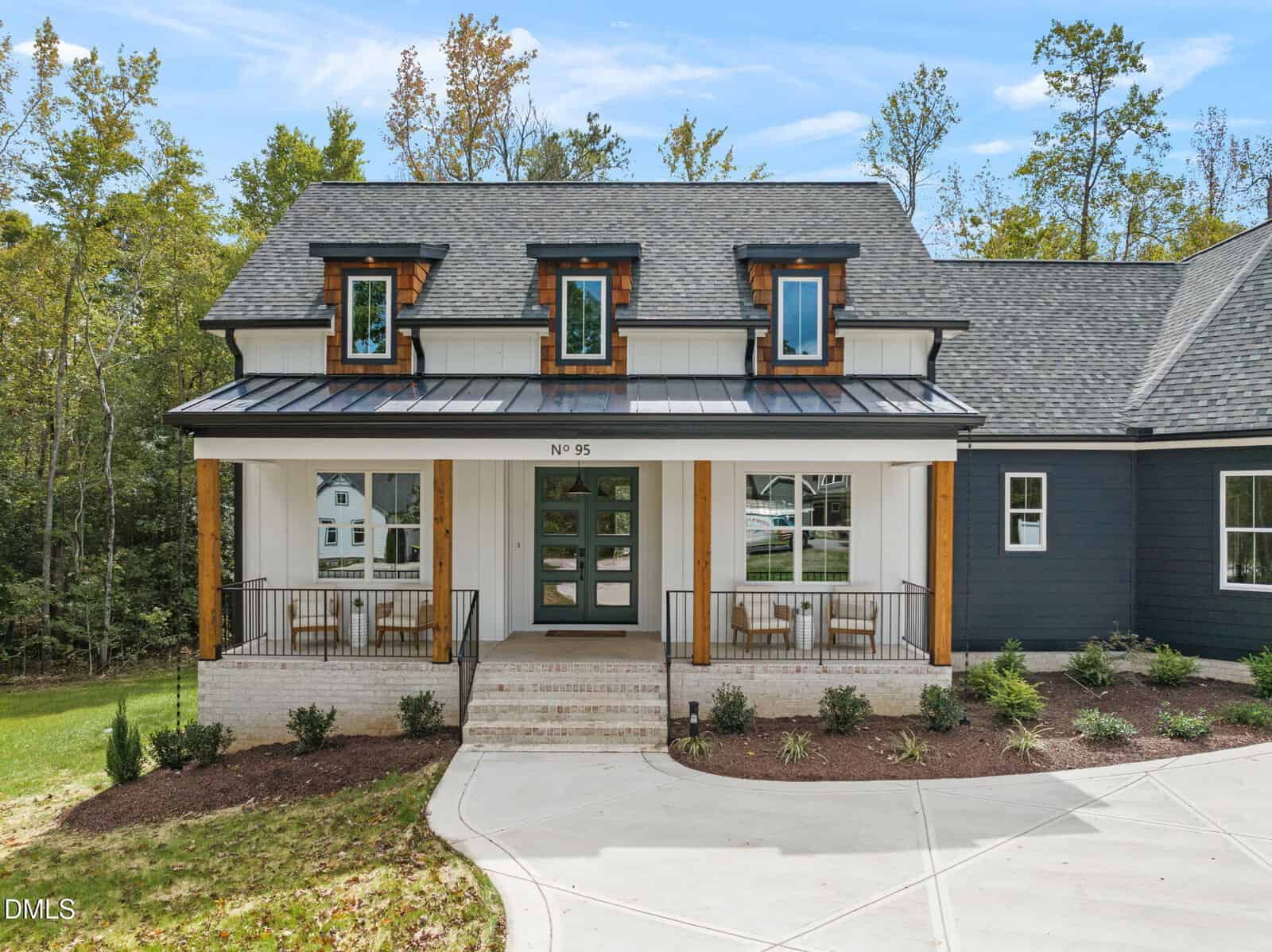 A modern two-story house with dark siding, white trim, three dormer windows, a covered front porch with chairs, and a wide front door. The yard has new landscaping, and trees are in the background.