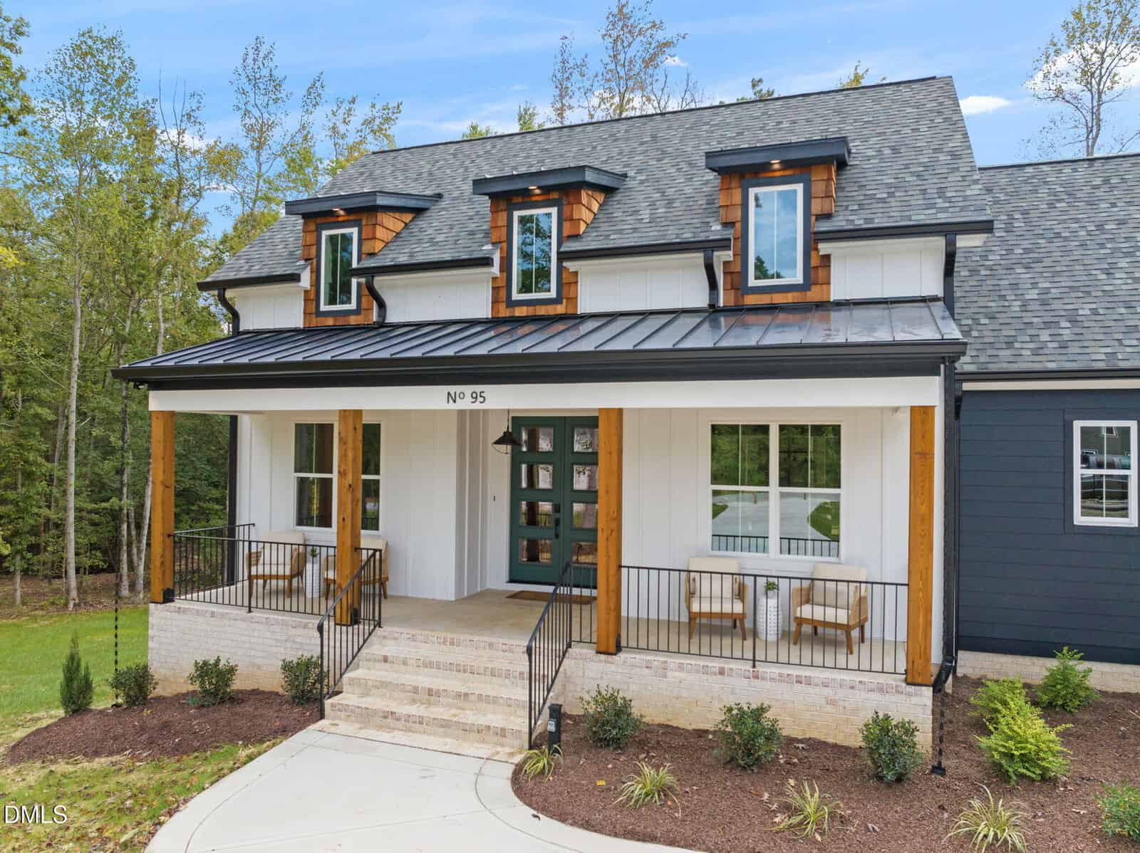 A modern two-story house with a large front porch, white siding, dark trim, and wood accents. There are three dormer windows on the roof and two chairs on the porch. Trees and shrubs surround the home.