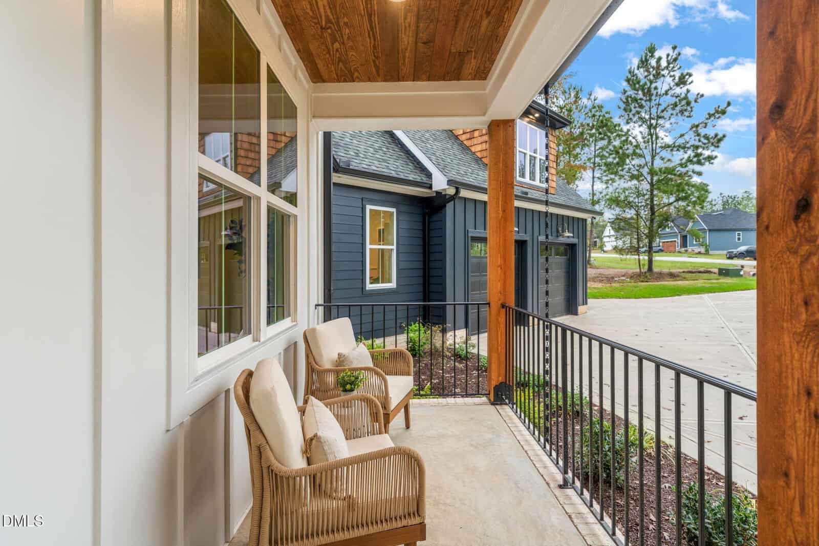 A cozy covered front porch with two wicker chairs and cushions overlooks a driveway and modern dark blue house exterior, with trees and a neighborhood visible in the background under a partly cloudy sky.