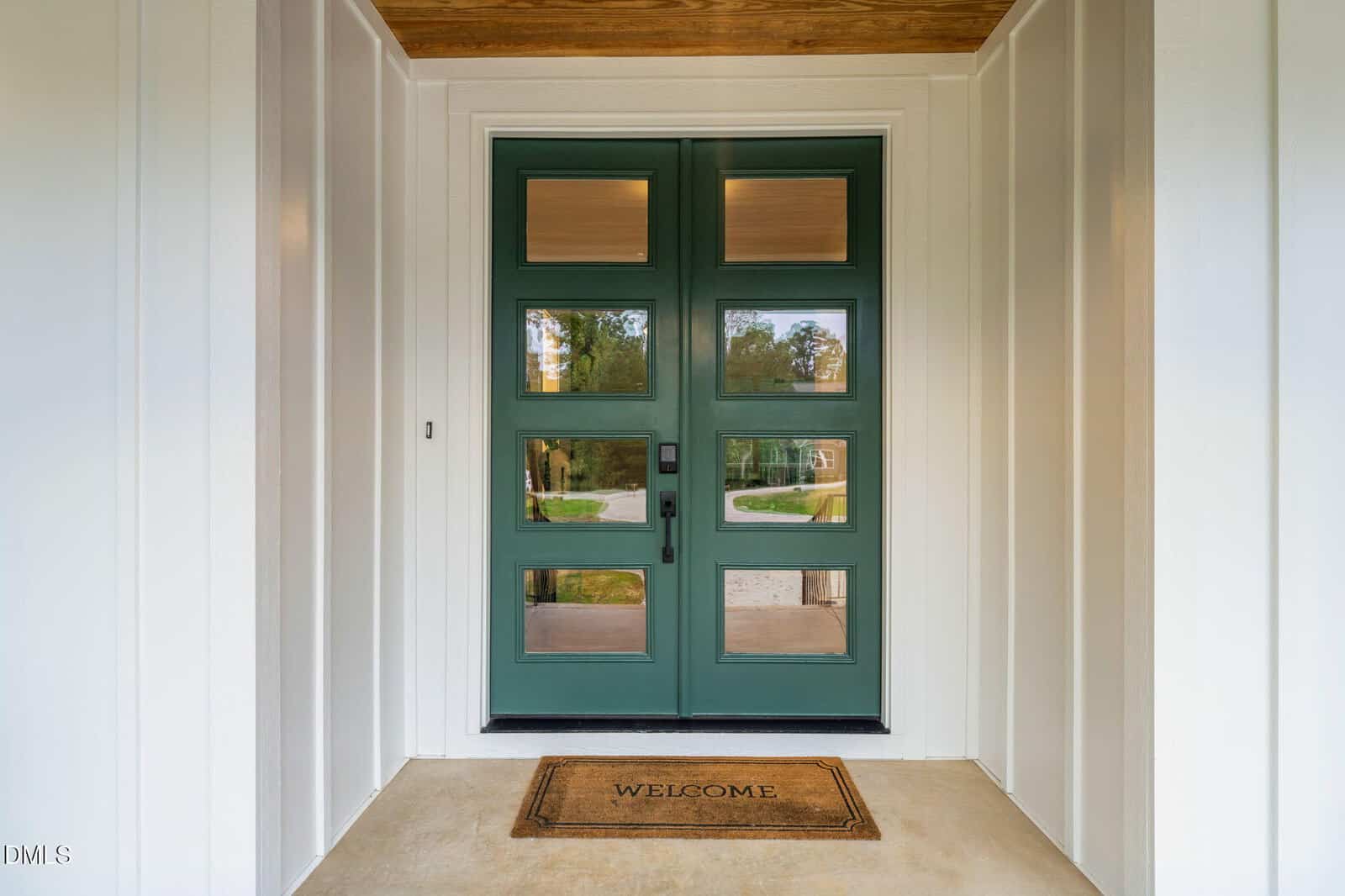 A green double front door with four glass panels on each side, set in a white-paneled entryway. A “Welcome” doormat is placed on the concrete porch floor in front of the doors. Trees and lawn are visible through the glass.