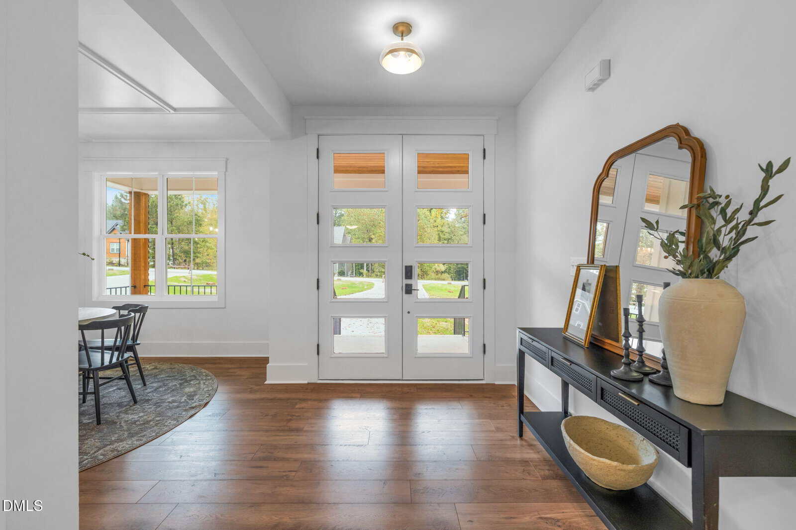 A bright entryway with white walls, wood flooring, a modern glass-paneled front door, a black console table holding a mirror, vase, basket, and framed photos, and a window showing a green yard outside.