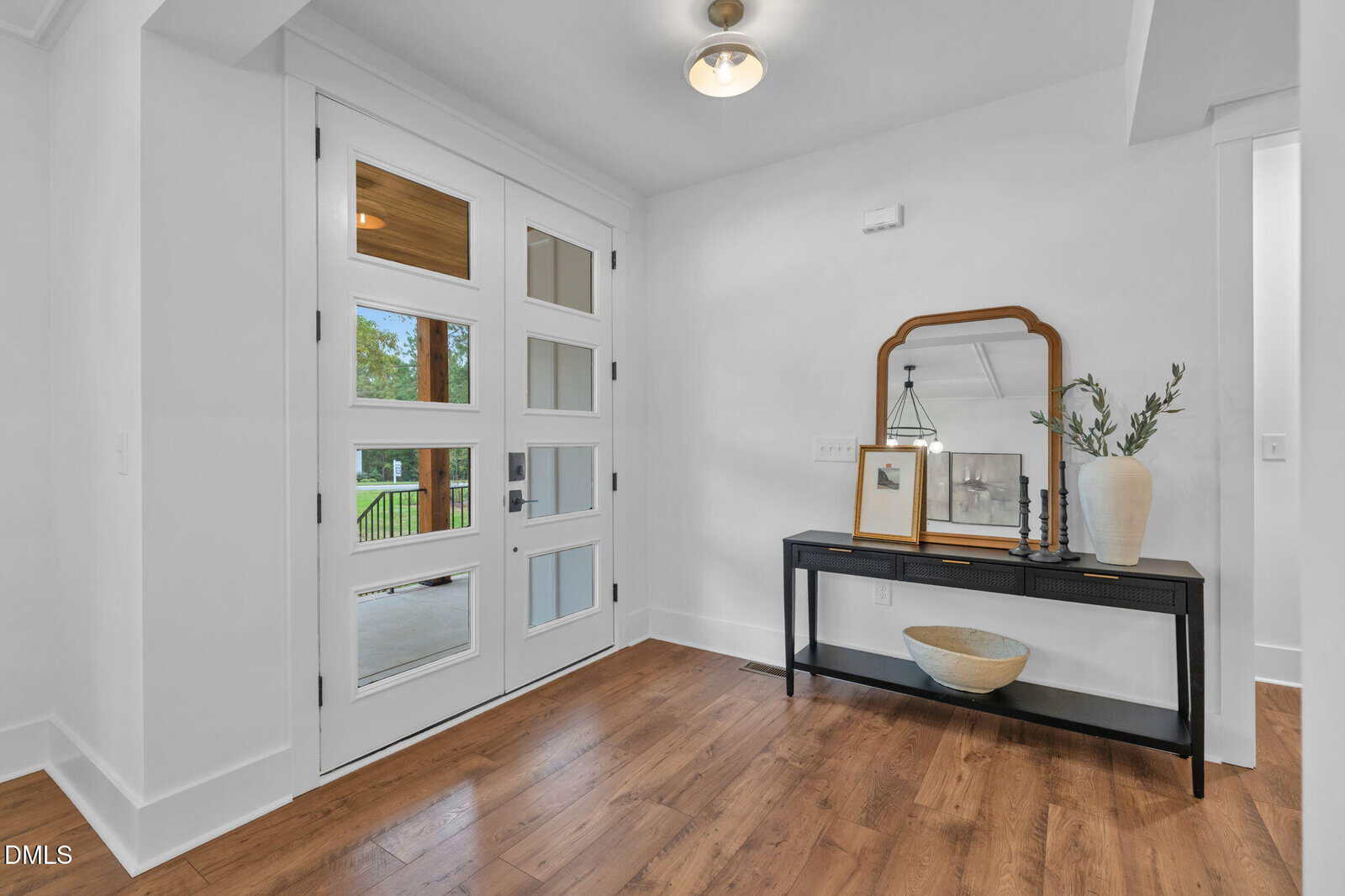 A modern entryway with a white front door featuring glass panels, wood flooring, and a black console table holding a mirror, lamp, vase with greenery, framed art, and a decorative bowl underneath.