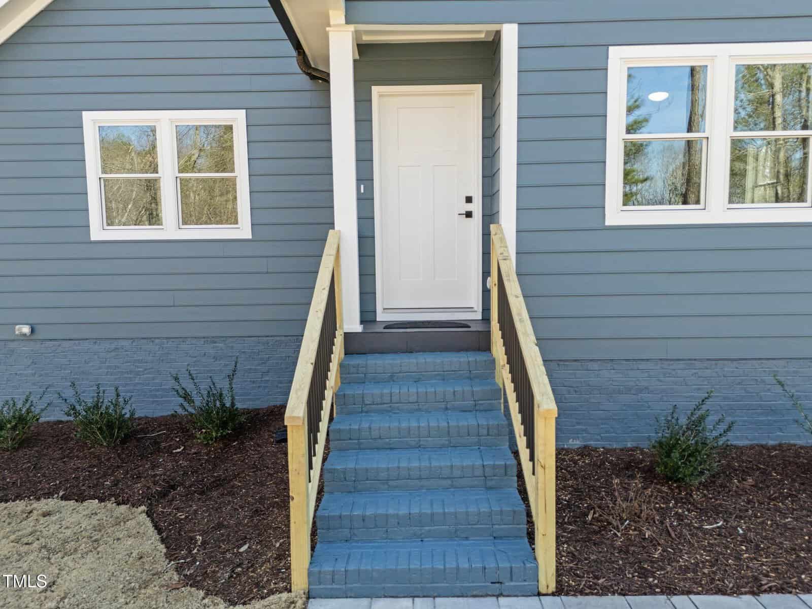 A blue house exterior with a white door. Six blue-painted brick steps with natural wood railings lead to the entrance. Two windows are visible on the right, with mulch and small green bushes lining the house.