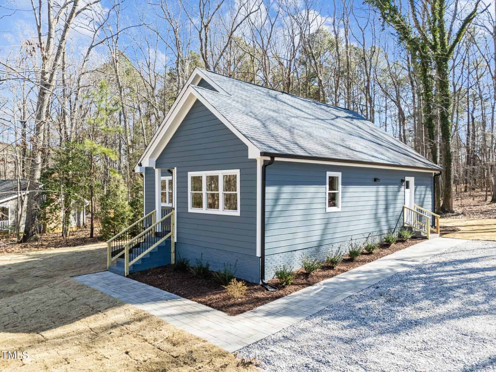 A small, blue single-story house with white trim, two entrances with steps, and a gravel driveway. The house is surrounded by bare trees and simple landscaping on a clear day.