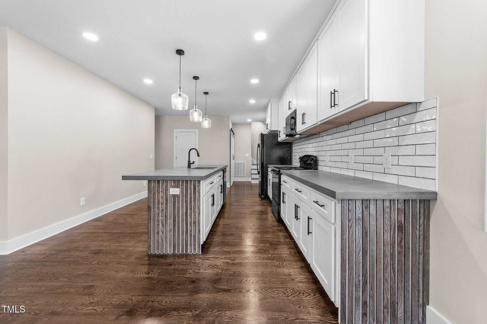 Modern kitchen with wood floors, gray countertops, white cabinets, stainless steel appliances, and two pendant lights over an island. Subway tile backsplash runs along the wall behind the counters.