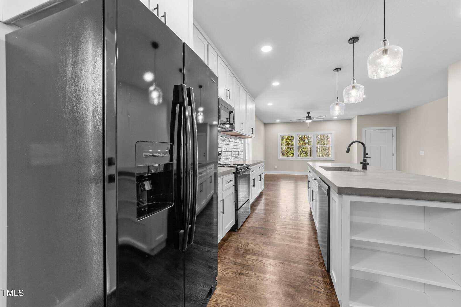 Modern kitchen with black refrigerator, white cabinets, stainless steel appliances, island with built-in shelves, pendant lights, and hardwood floors; large windows and ceiling fan visible in the background.