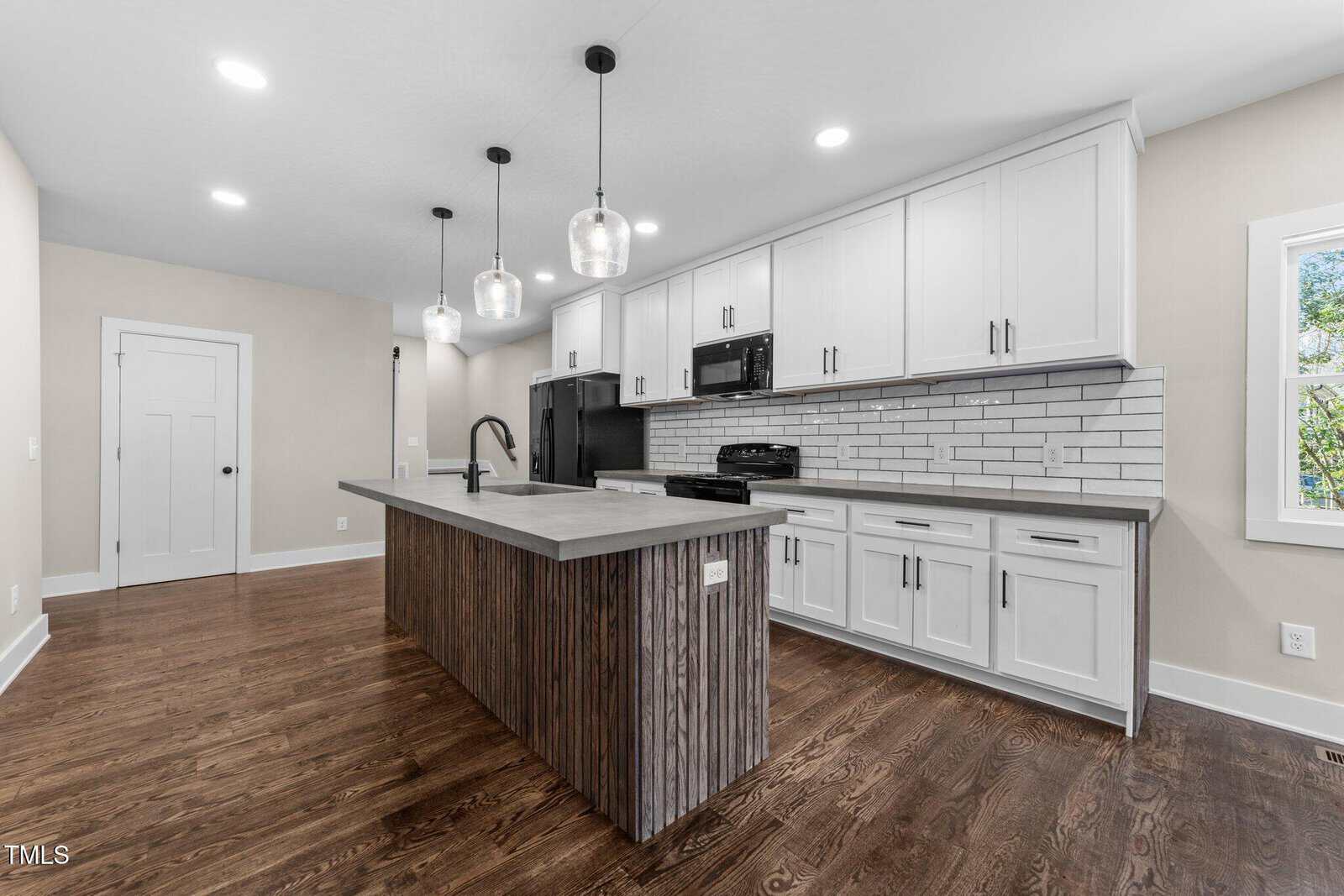 Modern kitchen with white cabinets, subway tile backsplash, a large island with dark wood accents, stainless steel appliances, pendant lighting, and hardwood floors. Natural light comes in through a window on the right.