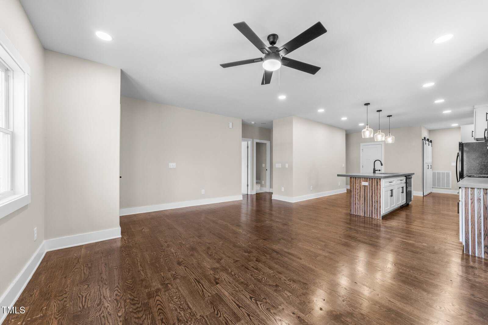 Spacious modern open-concept kitchen and living area with hardwood floors, beige walls, ceiling fan, recessed lighting, kitchen island with sink, and pendant lights. Large windows provide natural light.