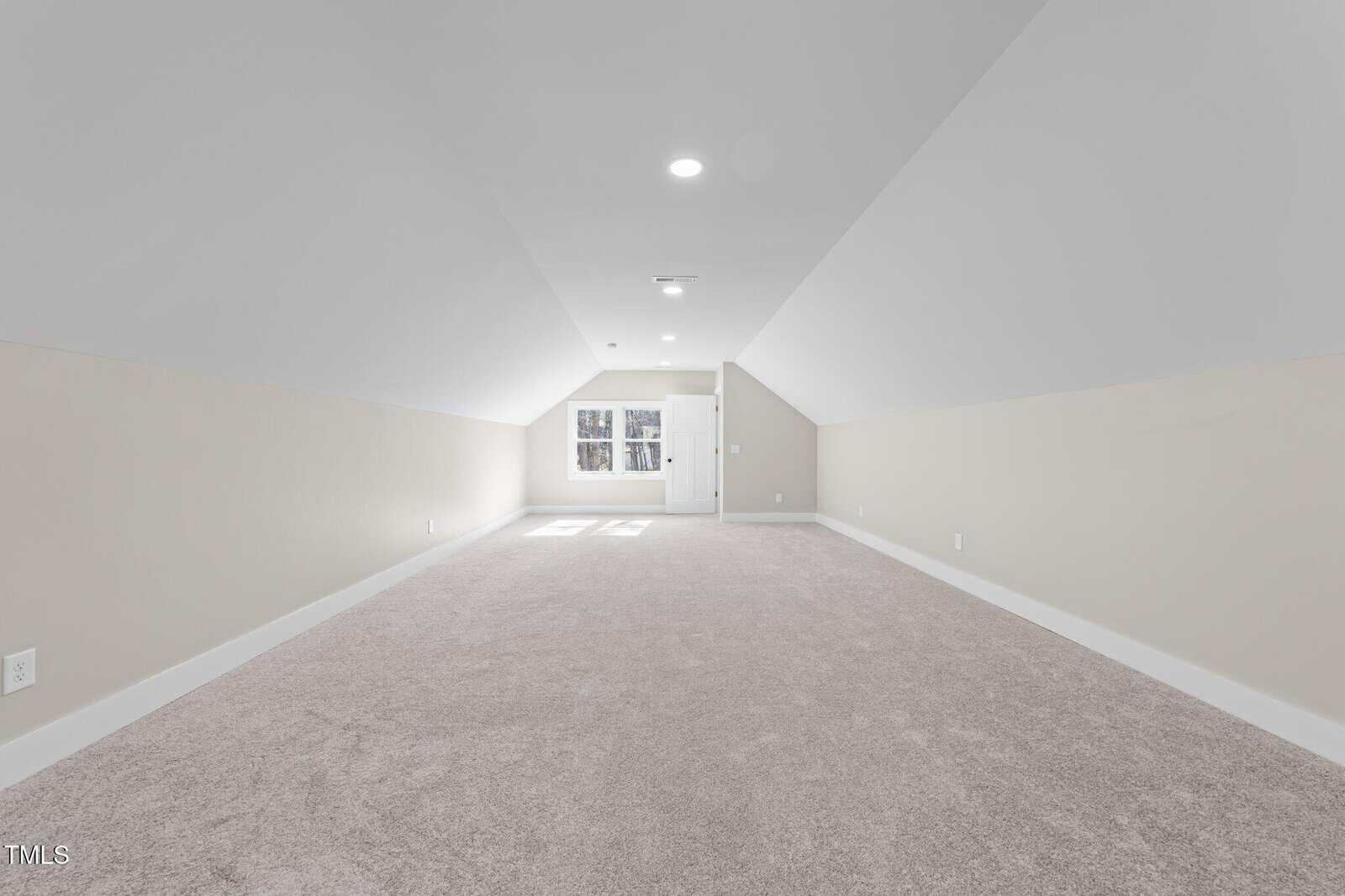 A spacious, empty attic room with light beige carpet, sloped white ceilings, and neutral walls. A window and door are at the far end, allowing natural light to brighten the clean, open space.