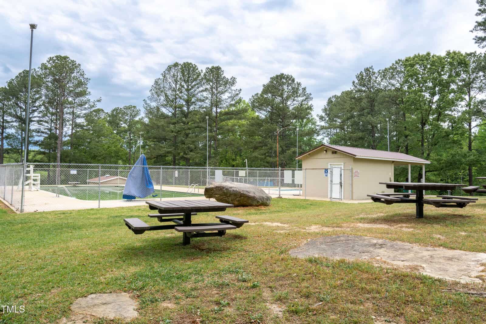 Fenced outdoor pool area with a blue tarp, nearby small building, picnic tables, and large rock on grassy ground, surrounded by trees under a cloudy sky.