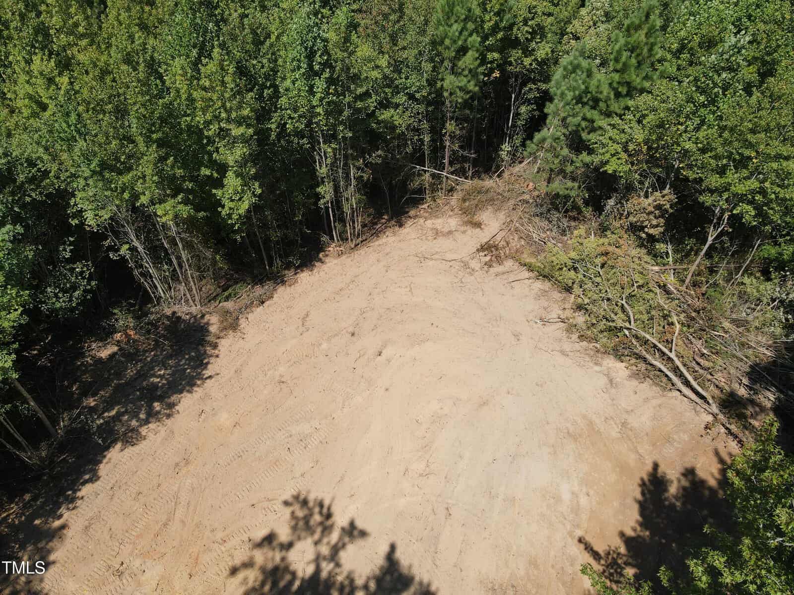 A cleared dirt area surrounded by dense green trees, with some fallen branches and tire tracks visible on the ground. The scene appears to be part of a construction or land-clearing project in a forested area.