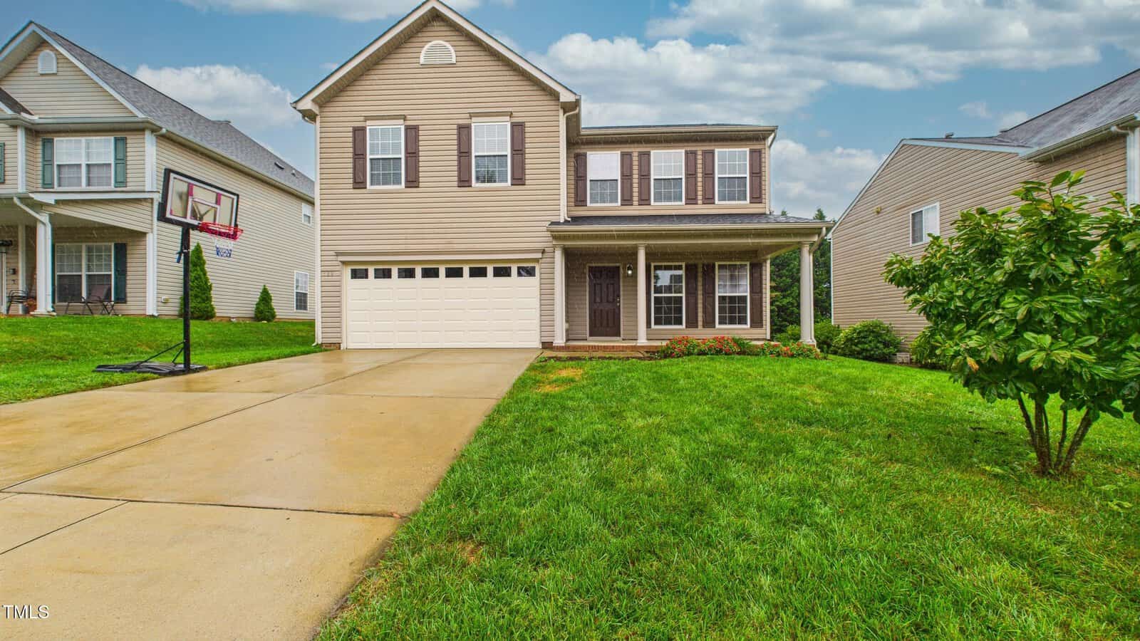 Two-story beige house with white trim, attached two-car garage, and front porch. Concrete driveway with a basketball hoop, green lawn, and neighboring houses on both sides under a partly cloudy sky.