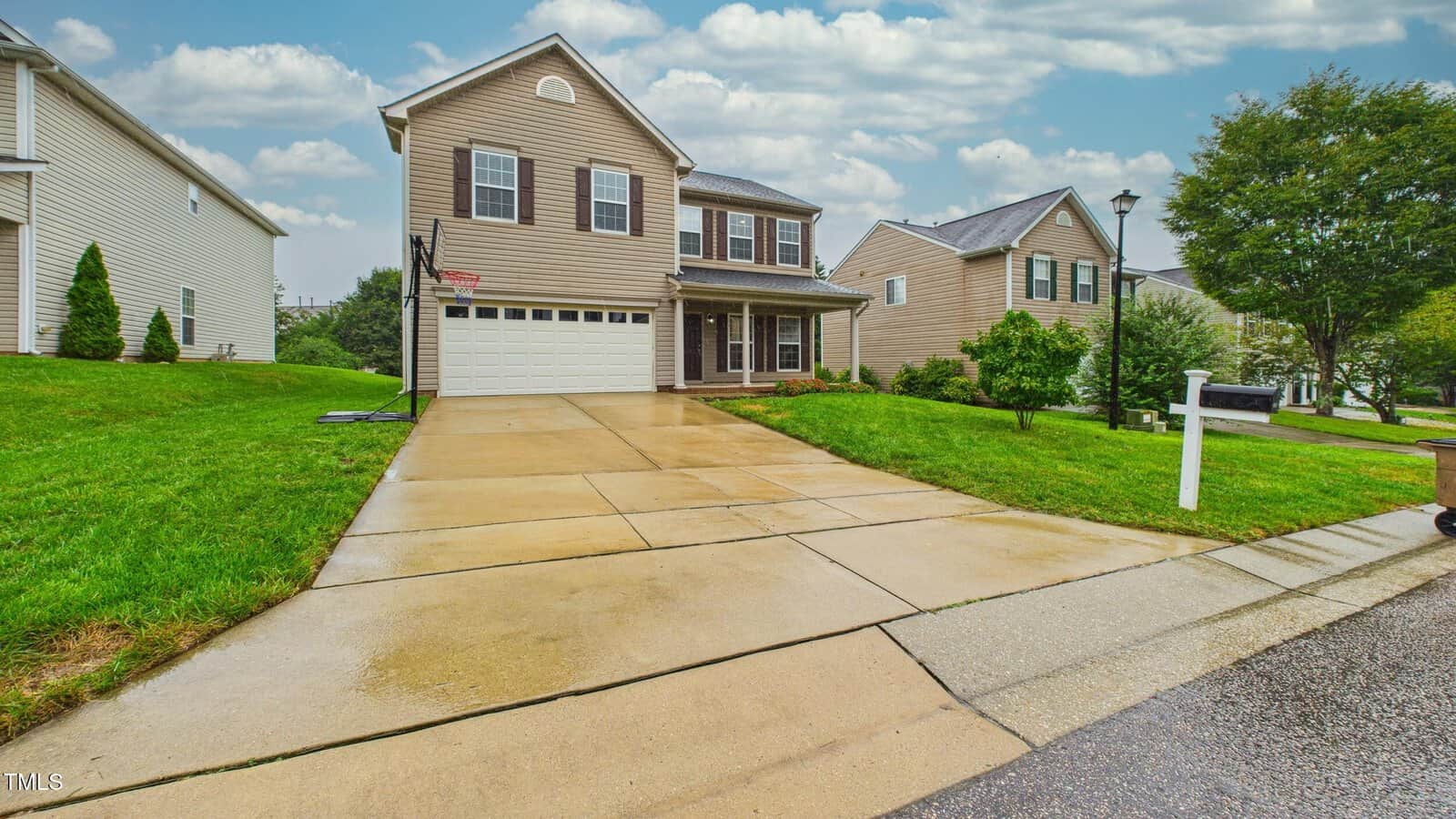 A two-story beige suburban house with a double garage, wet concrete driveway, green lawn, and surrounding houses, pictured on a cloudy day.