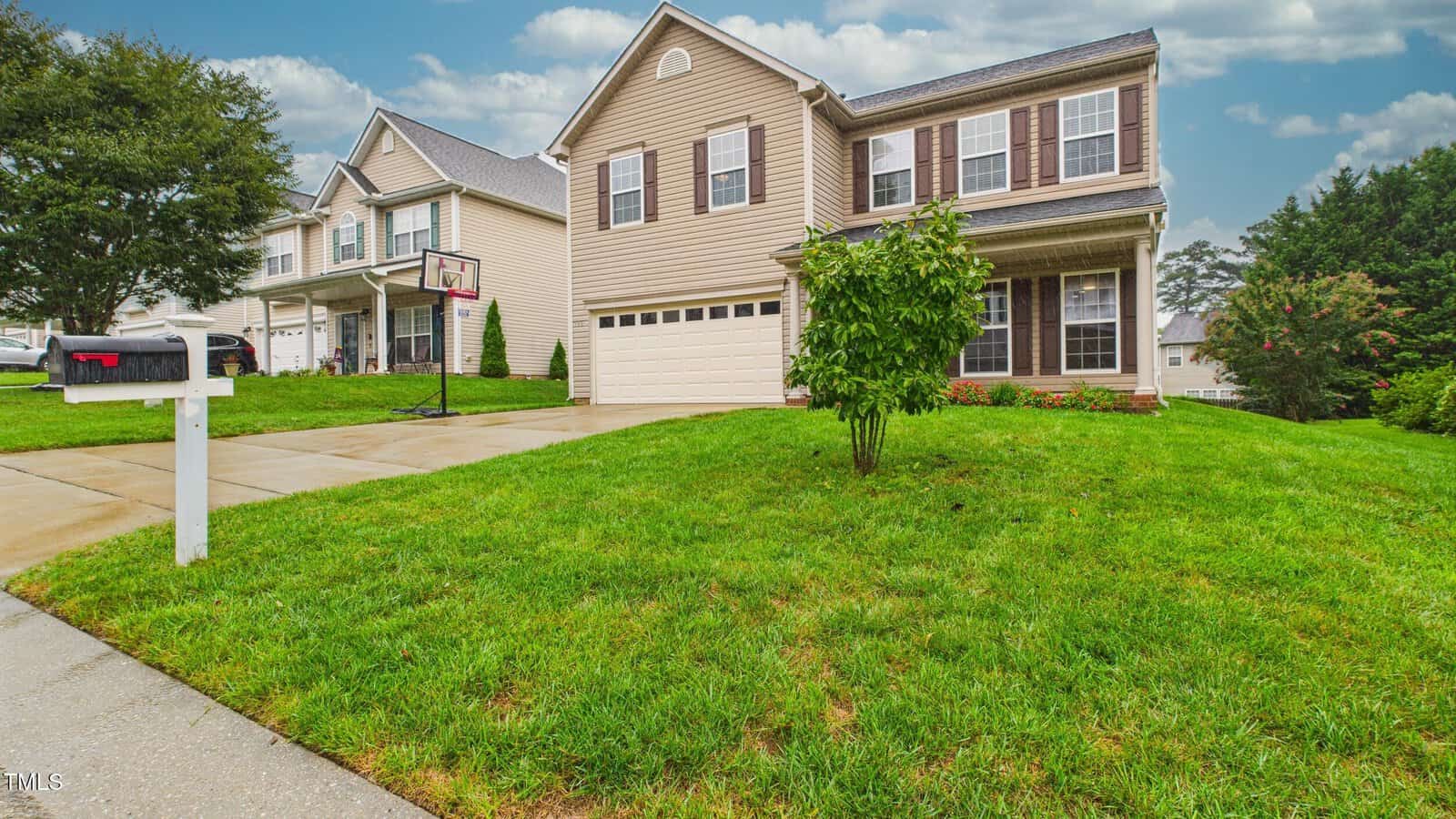 Two-story beige house with brown shutters, attached garage, and a small front porch. Green lawn with a tree, basketball hoop by the driveway, and a white mailbox, on a cloudy day in a suburban neighborhood.
