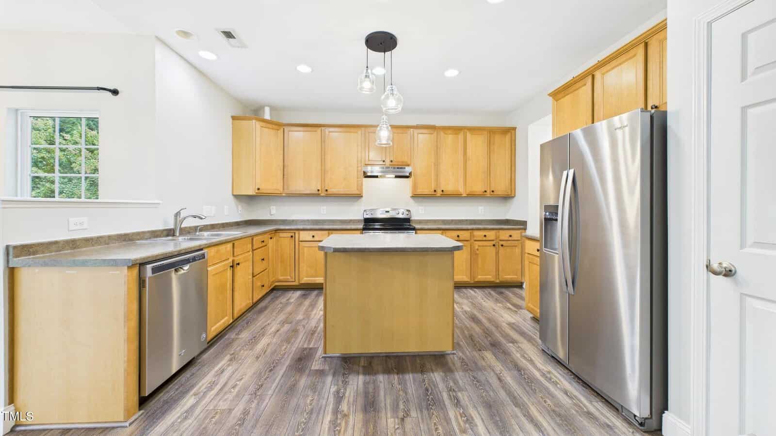 Bright, modern kitchen with light wood cabinets, stainless steel appliances, a small center island, gray countertops, wood-look flooring, and pendant lighting above the island. A window is above the sink on the left.